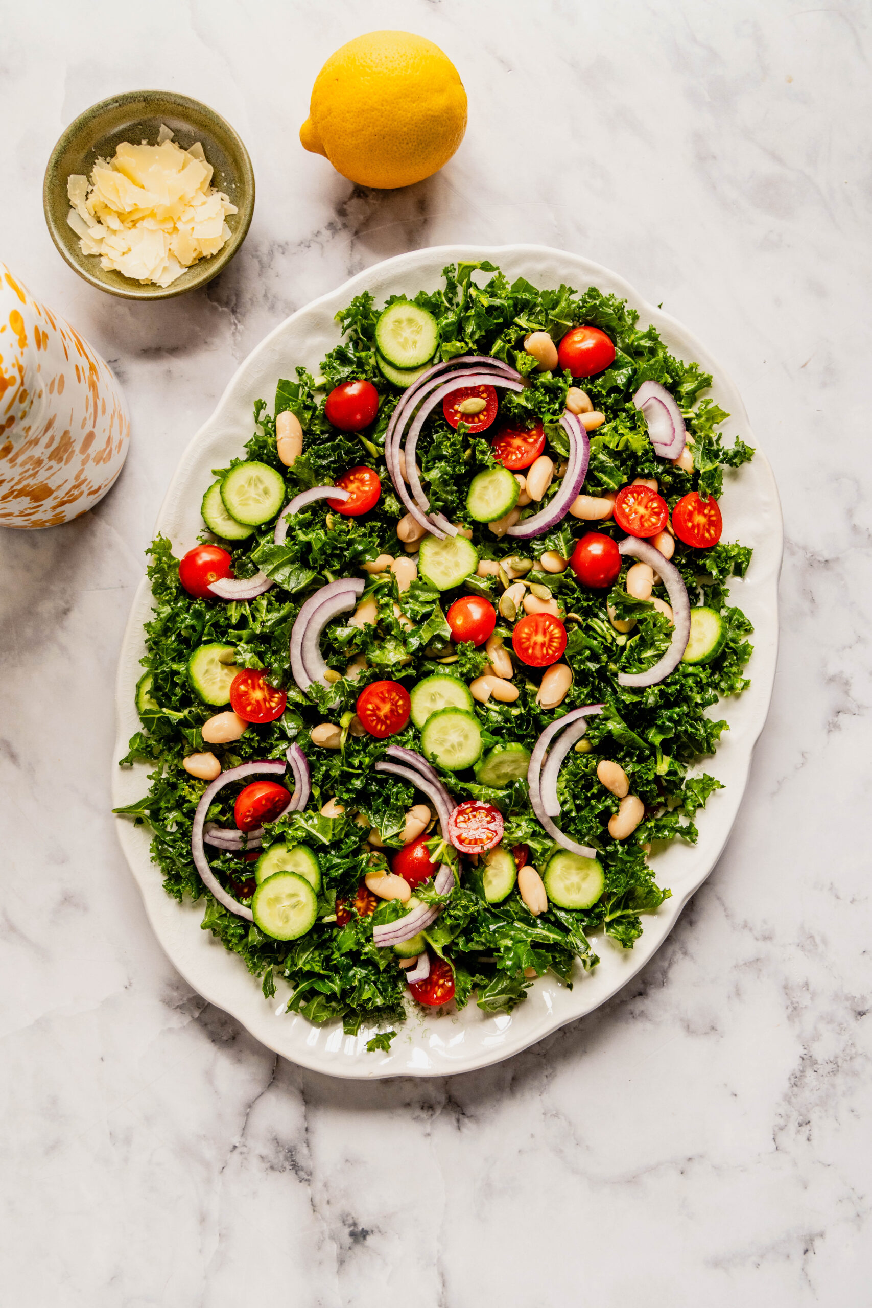 Overhead view of a kale and cannellini bean salad on a white serving platter, topped with sliced Persian cucumbers, halved grape tomatoes, thin red onion slices, and pepitas, with shaved Pecorino and a lemon on a marble countertop nearby.