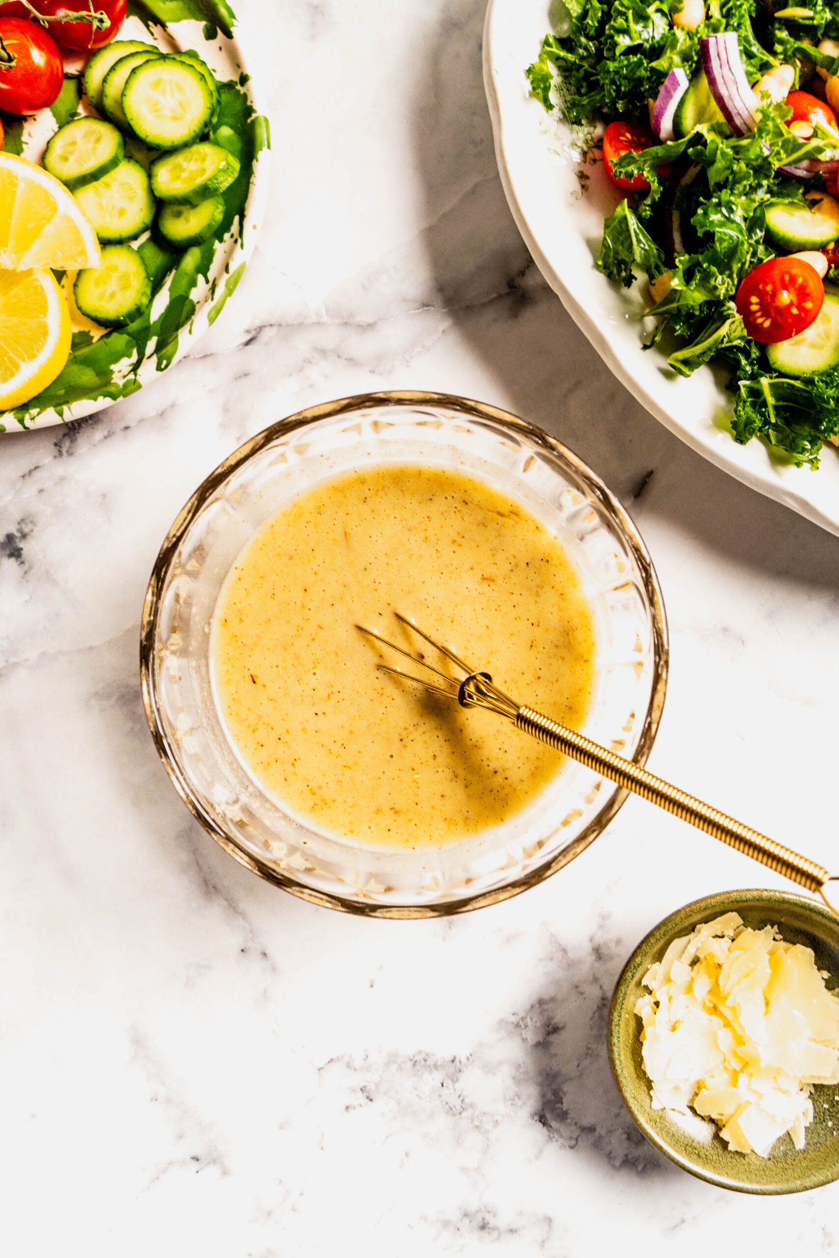 Overhead view of lemon Dijon vinaigrette being whisked in a glass bowl for kale and cannellini bean salad, with sliced cucumbers, cherry tomatoes, chopped kale, and shaved Pecorino nearby on a white marble surface.