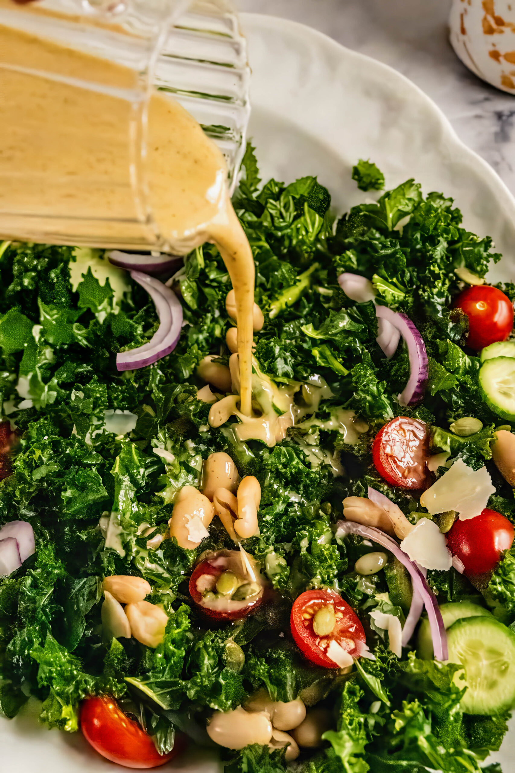 Close-up of lemon Dijon dressing being poured over kale and cannellini bean salad with cherry tomatoes, sliced cucumbers, red onion, pepitas, and shaved Pecorino on a white platter.
