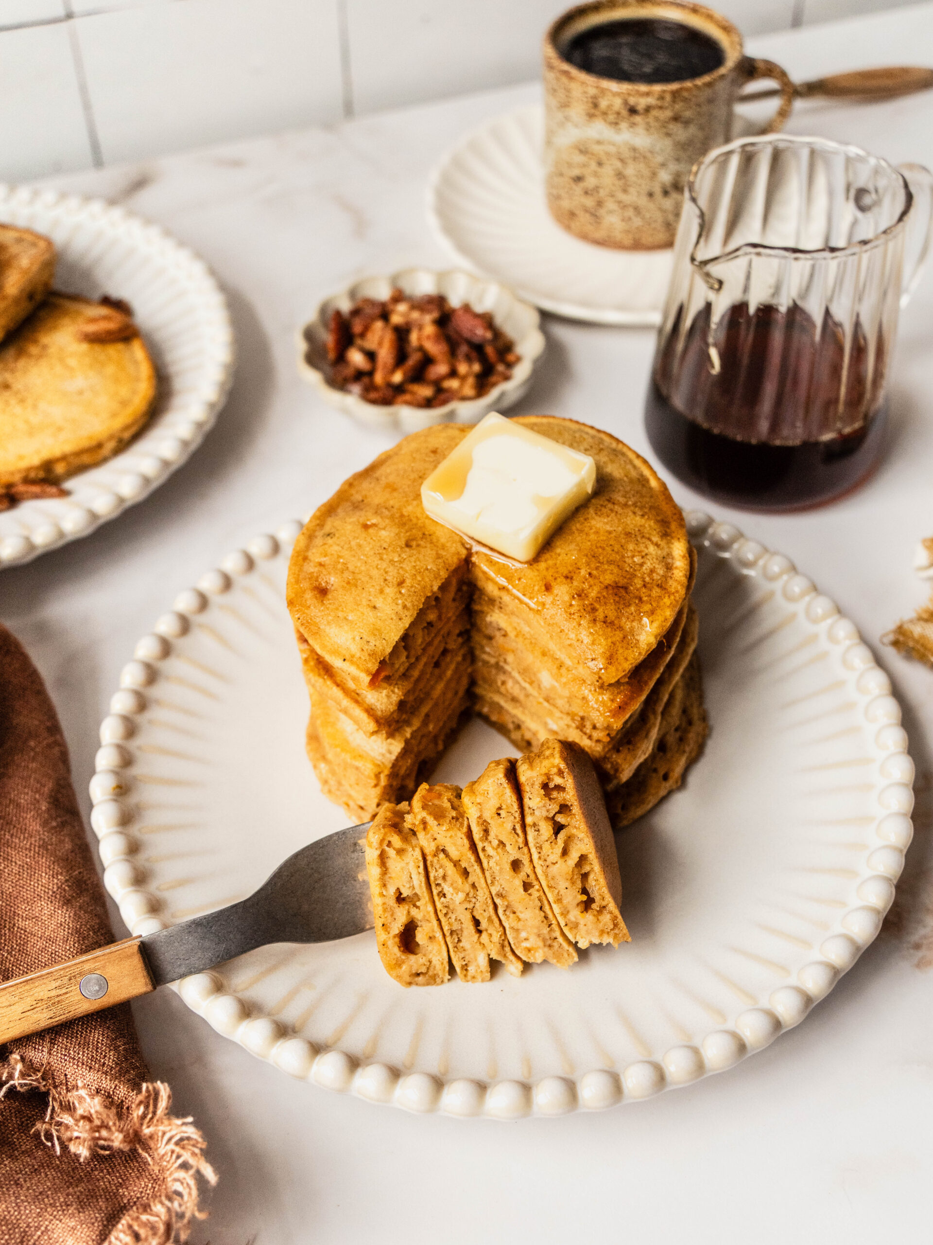 Gluten free sweet potato pancakes stacked on a white scalloped plate, topped with a pat of butter and maple syrup, with a slice cut out to show the fluffy interior and a fork holding a bite. A glass syrup pitcher, pecans, and eggs sit on a white marble background in warm natural light.