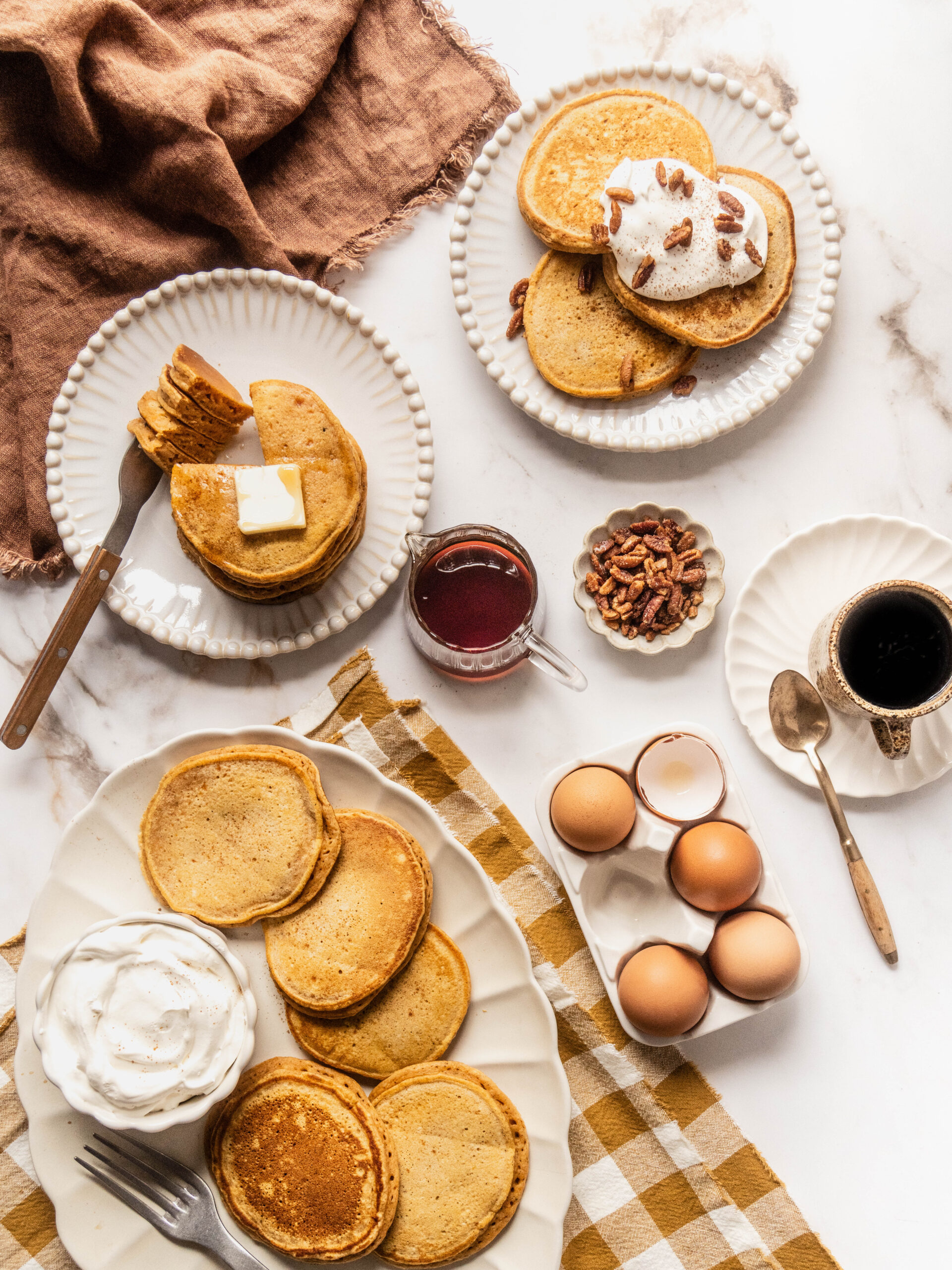 Overhead brunch spread of gluten free sweet potato pancakes served on white scalloped plates with butter, maple syrup, whipped cream, and candied pecans, styled on a white marble table with coffee and fresh eggs in warm natural light.