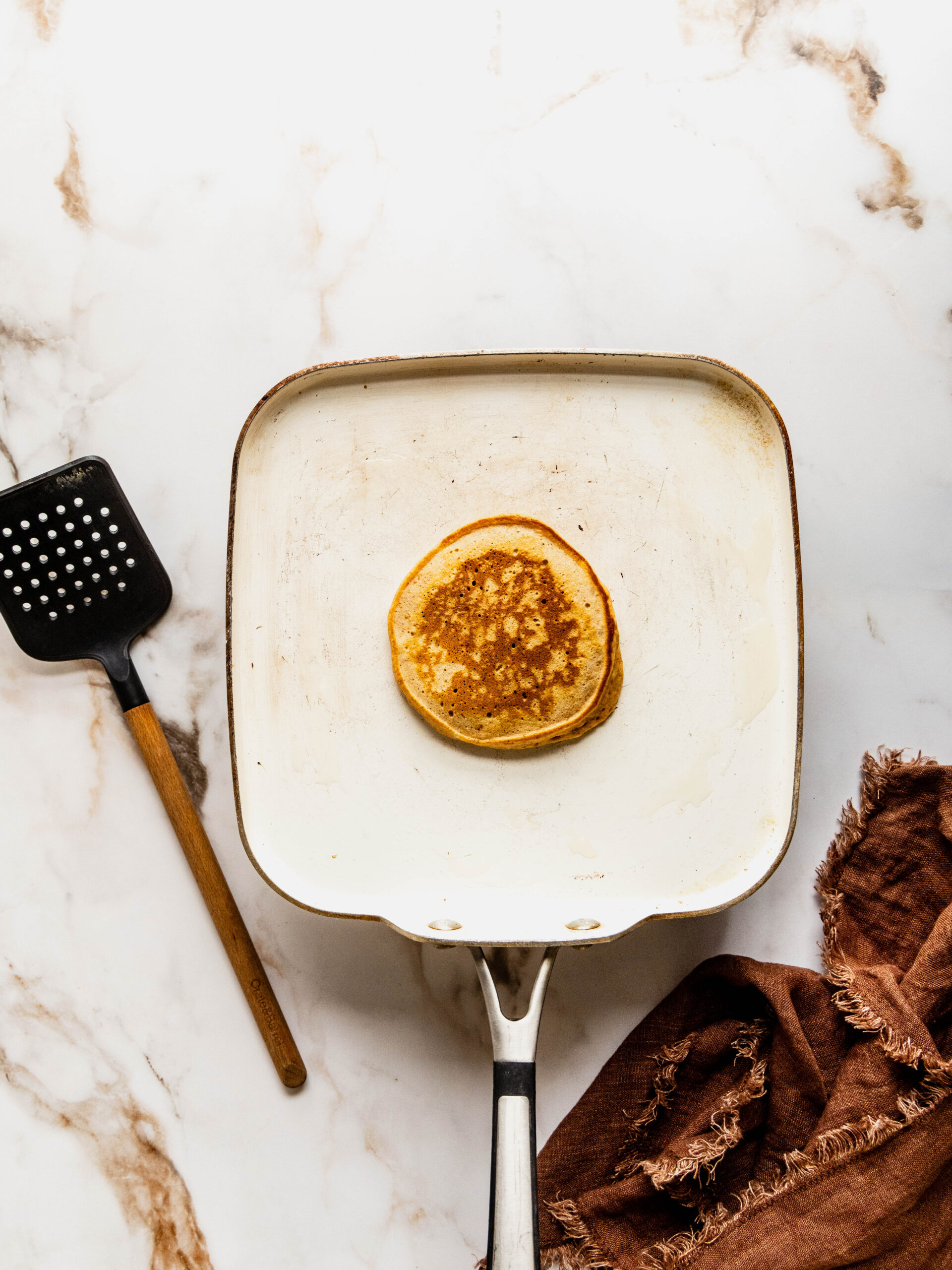 Overhead view of gluten free sweet potato pancakes cooking in a white skillet, showing a golden brown pancake with crisp edges, styled on a white marble countertop with a spatula and brown linen napkin nearby.