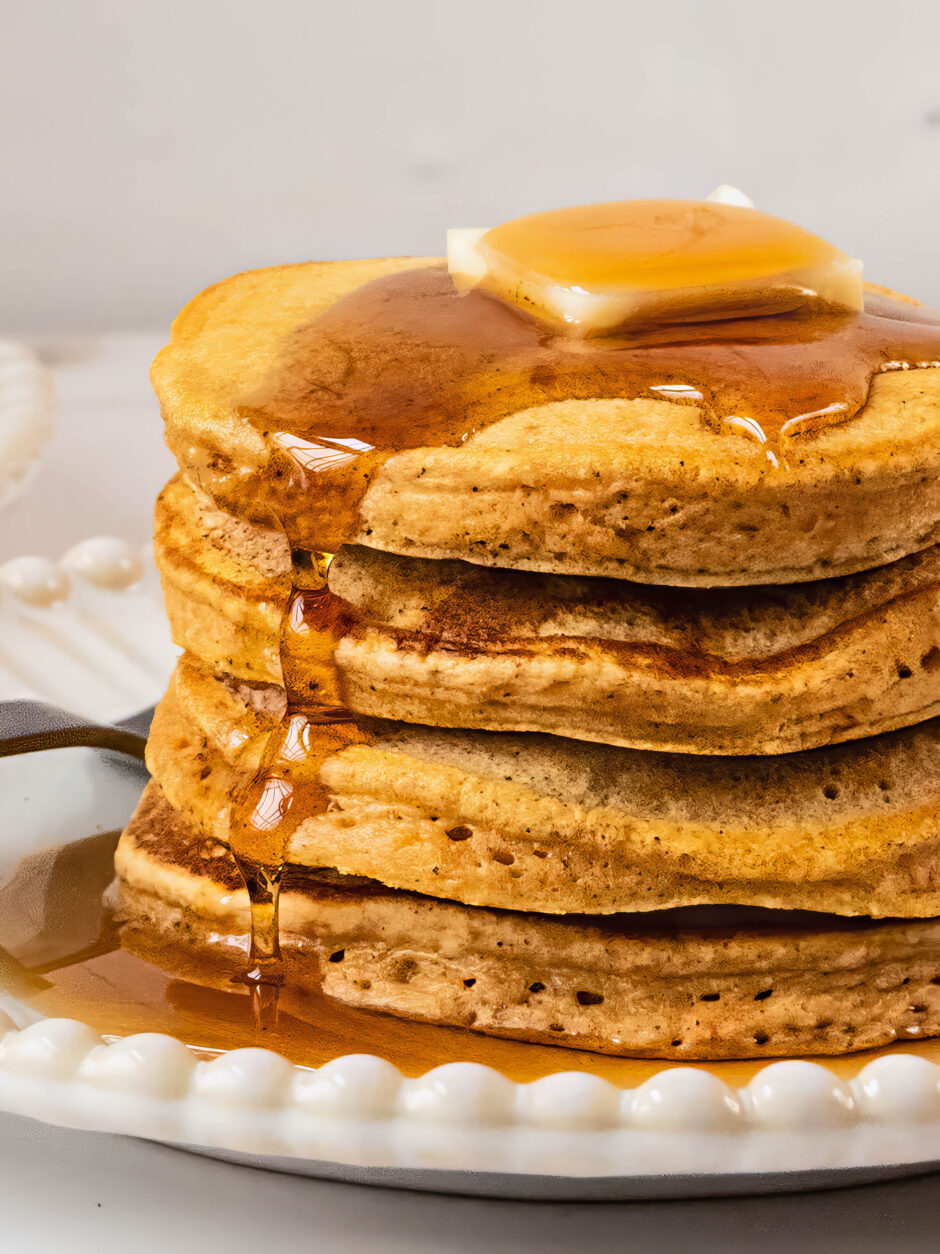 Close-up of gluten free sweet potato pancakes stacked high on a white plate, topped with a pat of melting butter and maple syrup dripping down the fluffy layers in warm natural light.