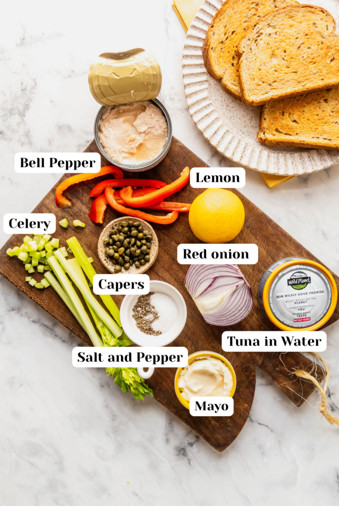 Overhead ingredient shot for caper tuna salad featuring canned tuna, capers, diced celery, red onion, sliced red bell pepper, lemon, mayonnaise, salt and pepper, with toasted bread on the side on a wooden board.