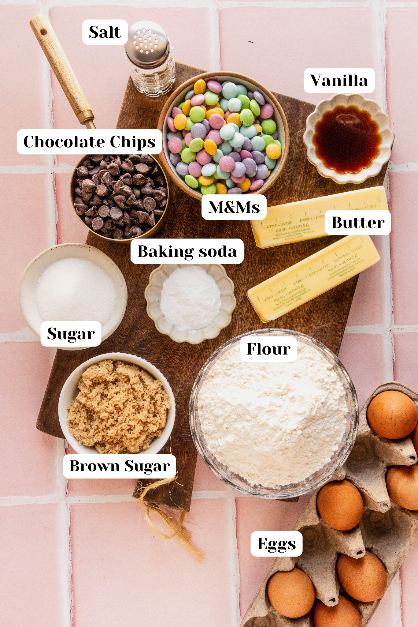 Bright overhead image of ingredients for Easter bunny cookies arranged on a wooden board over a pink tiled background, including flour, brown sugar, sugar, eggs, butter, vanilla, baking soda, chocolate chips, and pastel M&Ms in individual bowls.