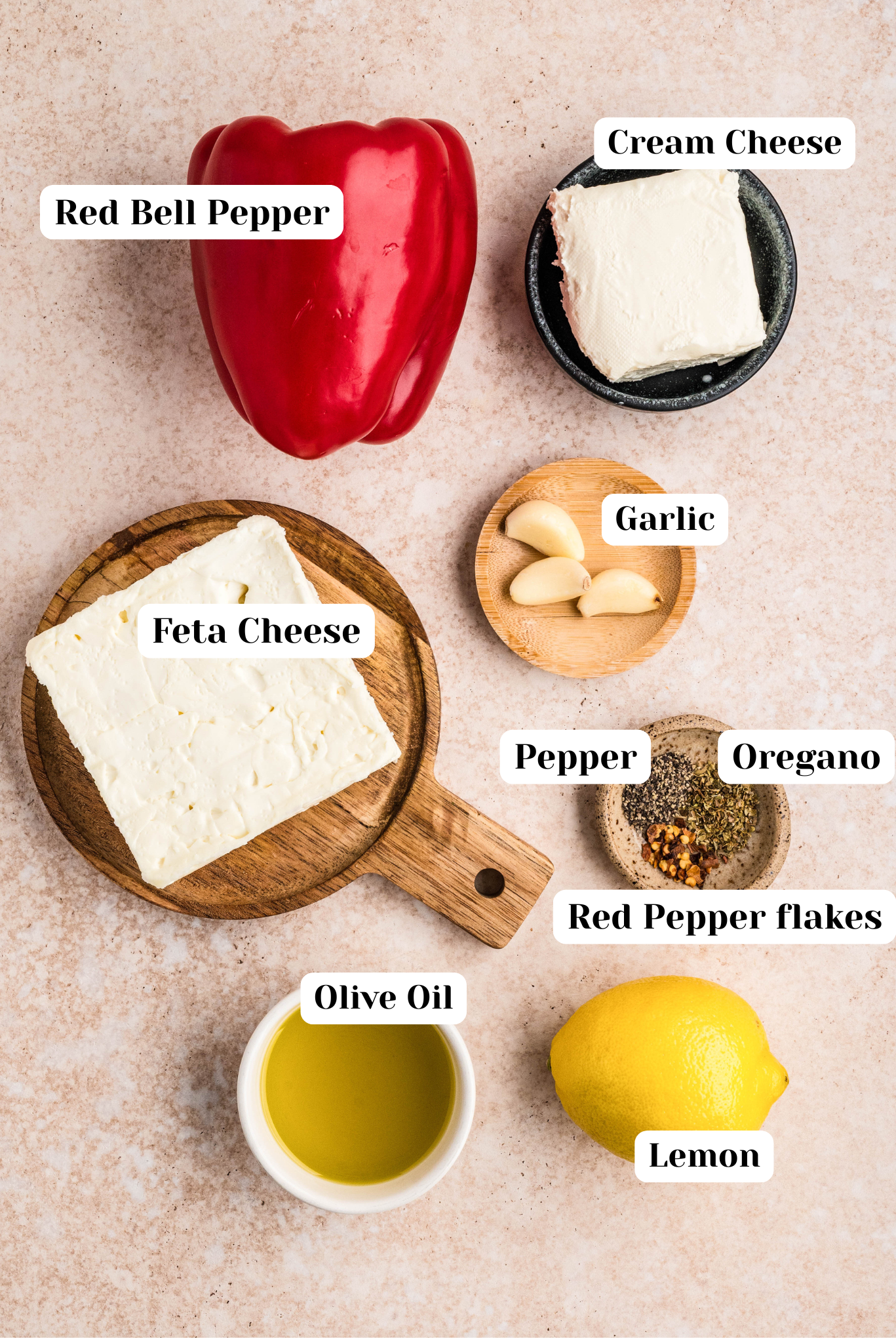 Overhead ingredient shot for feta red pepper dip featuring a whole red bell pepper, blocks of feta and cream cheese, fresh garlic cloves, dried oregano, cracked black pepper and red pepper flakes, a lemon, and a small bowl of olive oil arranged on a neutral stone surface.