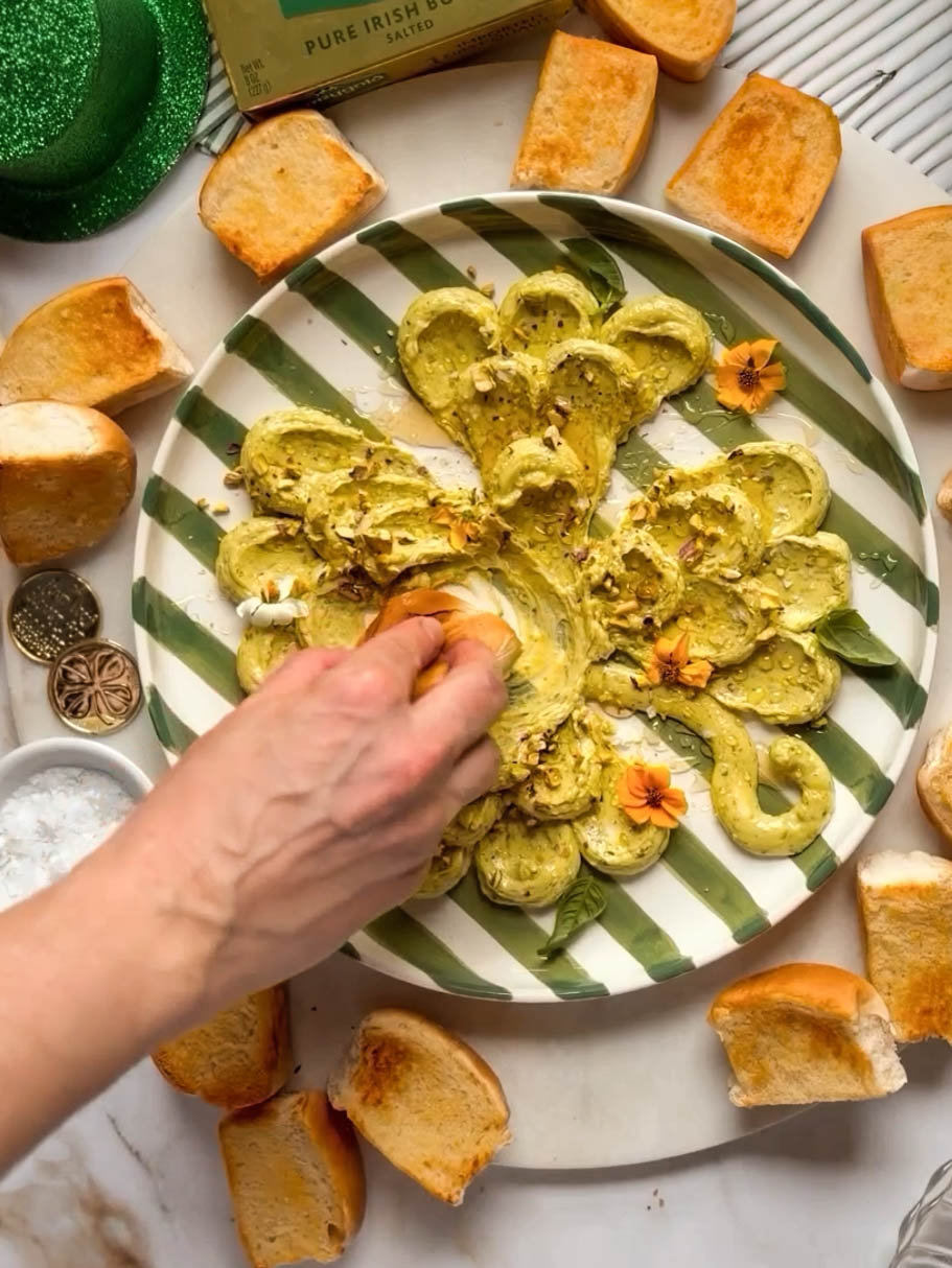 Hand dipping toasted bread into a shamrock-shaped pesto butter board topped with pistachios, honey, edible flowers, and basil, surrounded by toasted brioche rolls and butter on a marble surface.