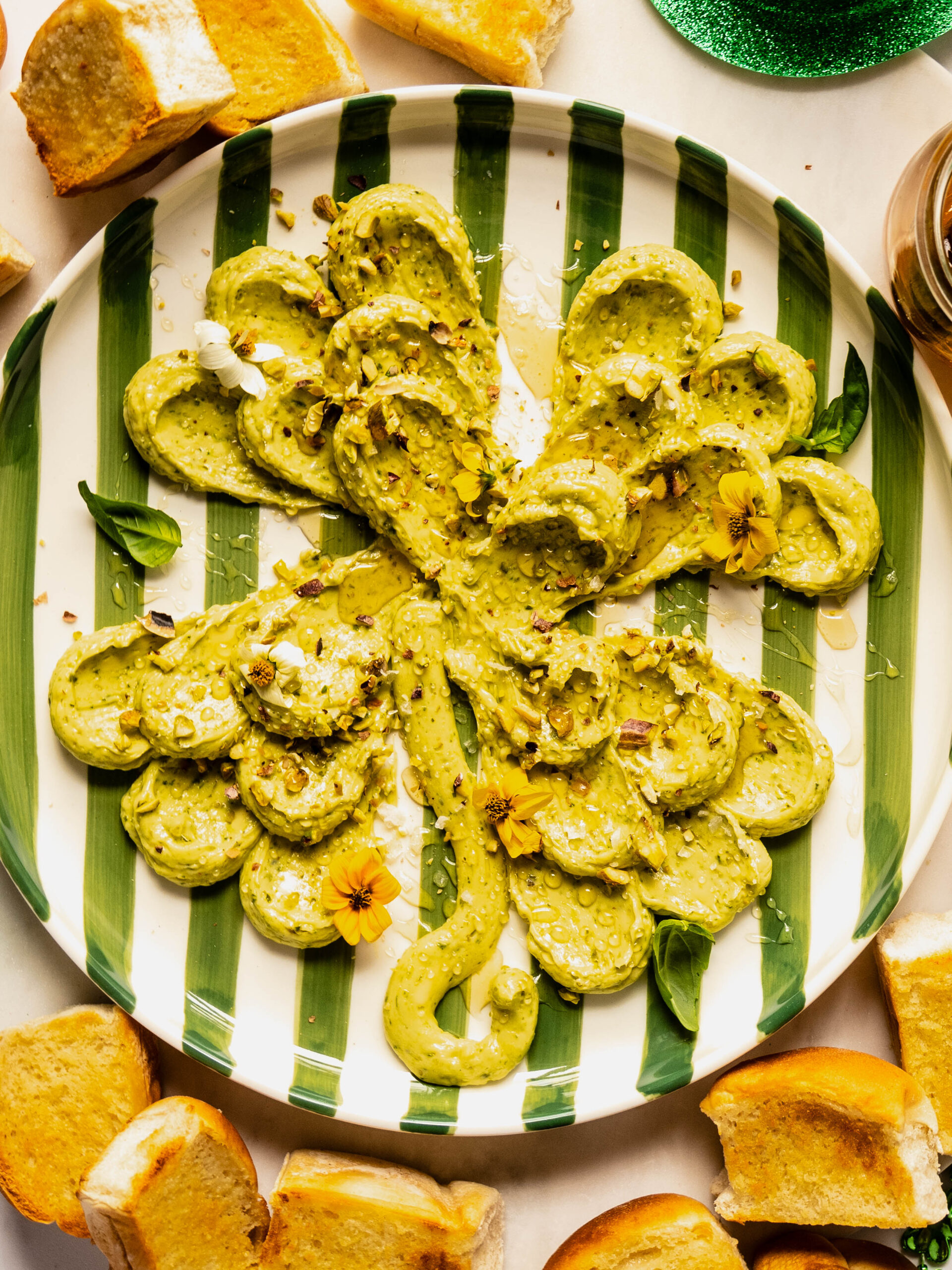 overhead shot of Shamrock-shaped pesto butter board topped with chopped pistachios, honey drizzle, edible flowers, and fresh basil, served on a striped plate with toasted bread slices for dipping.