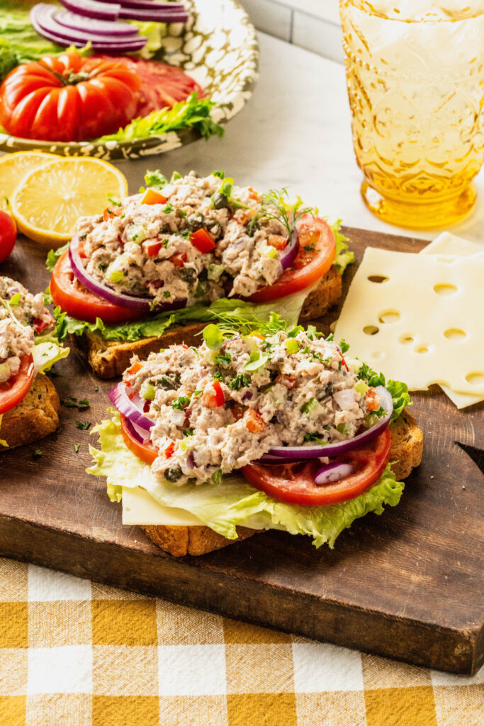 Side view of caper tuna salad piled onto toasted bread with layers of lettuce, tomato, red onion, and cheese, garnished with fresh herbs and capers, served on a rustic wooden board with lemon and cherry tomatoes nearby.