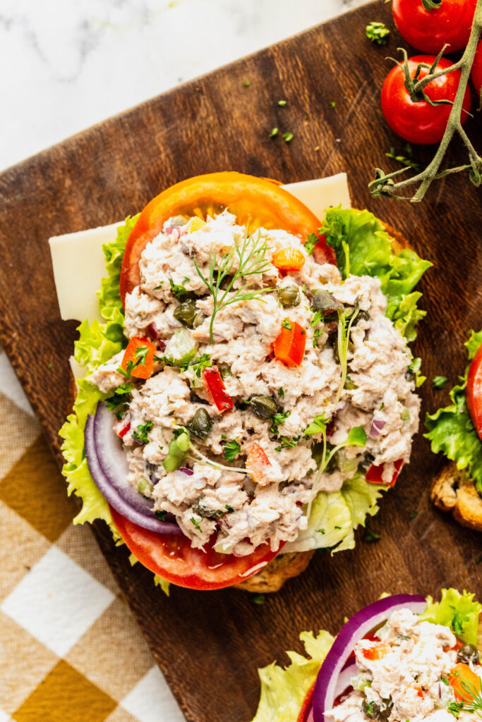 Overhead view of caper tuna salad piled onto toasted bread with layers of lettuce, tomato, red onion, and cheese, garnished with fresh herbs and capers, served on a rustic wooden board with lemon and cherry tomatoes nearby.