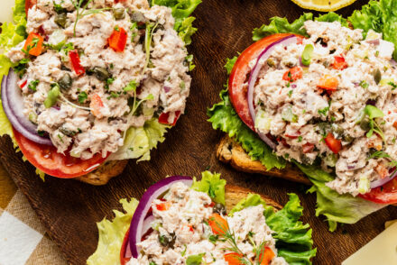 Bright overhead shot of caper tuna salad piled onto toasted bread with crisp lettuce, sliced tomatoes, and red onion, garnished with fresh herbs and capers, styled on a wooden board with lemon slices and cherry tomatoes.