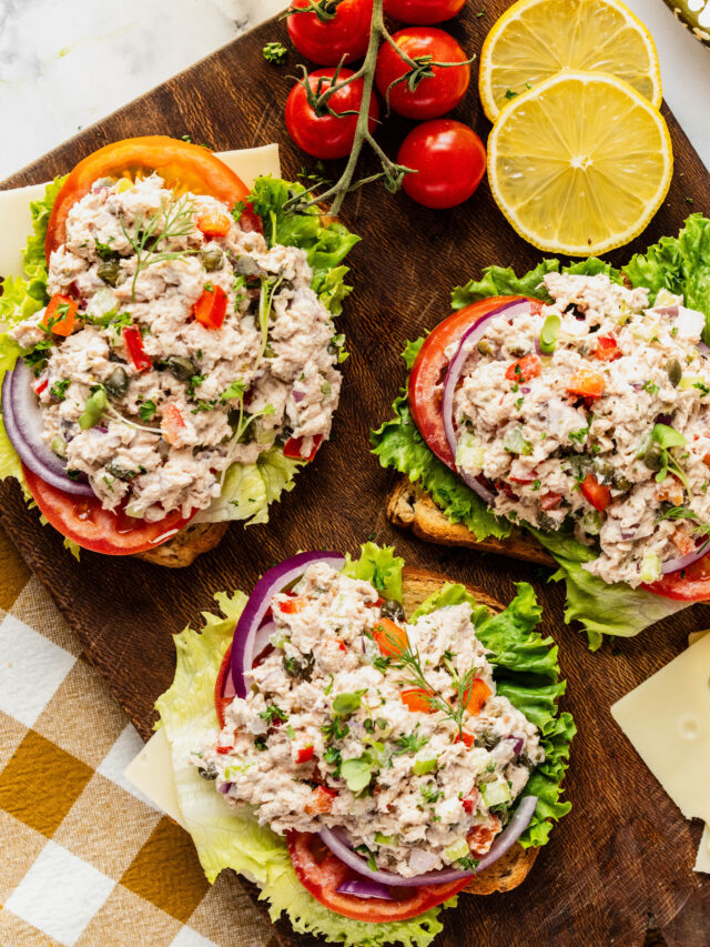 Bright overhead shot of caper tuna salad piled onto toasted bread with crisp lettuce, sliced tomatoes, and red onion, garnished with fresh herbs and capers, styled on a wooden board with lemon slices and cherry tomatoes.