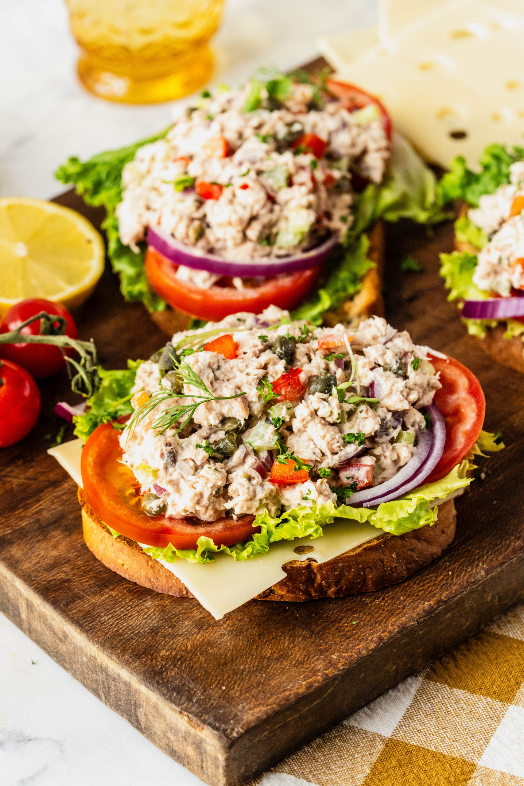 Side view of caper tuna salad piled onto toasted bread with layers of lettuce, tomato, red onion, and cheese, garnished with fresh herbs and capers, served on a rustic wooden board with lemon and cherry tomatoes nearby.