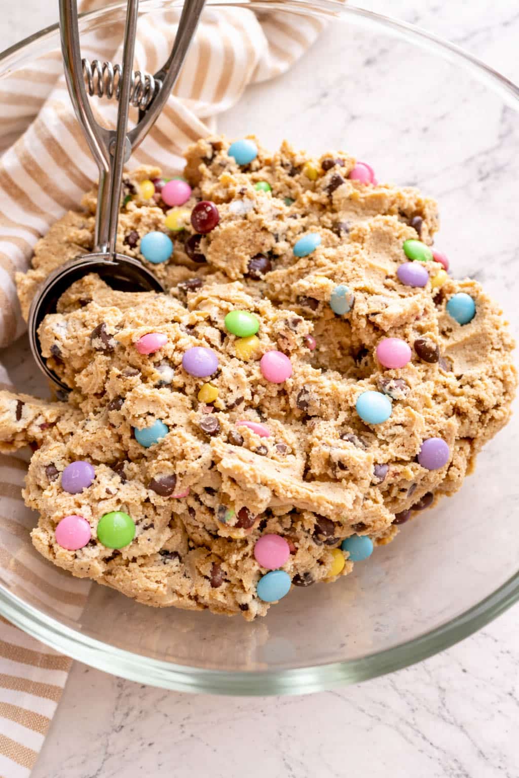 Close-up of Easter bunny cookie dough in a glass bowl, mixed with pastel M&Ms and chocolate chips, with a cookie scoop resting beside the soft, colorful dough.