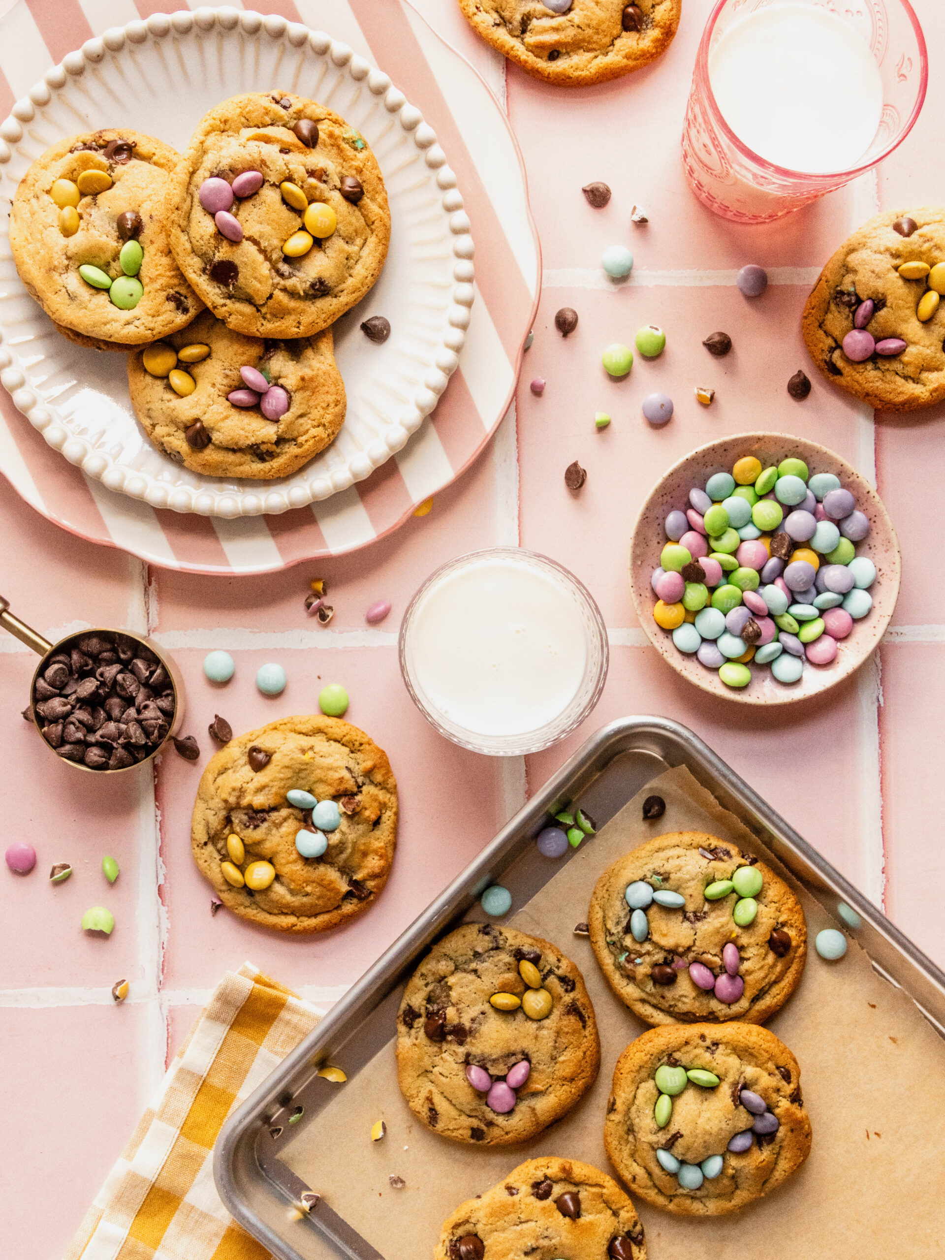 Bright overhead spread of Easter bunny cookies decorated with pastel M&Ms and chocolate chips, arranged on plates and a baking sheet with scattered candies and a glass of milk on a pink spring background.