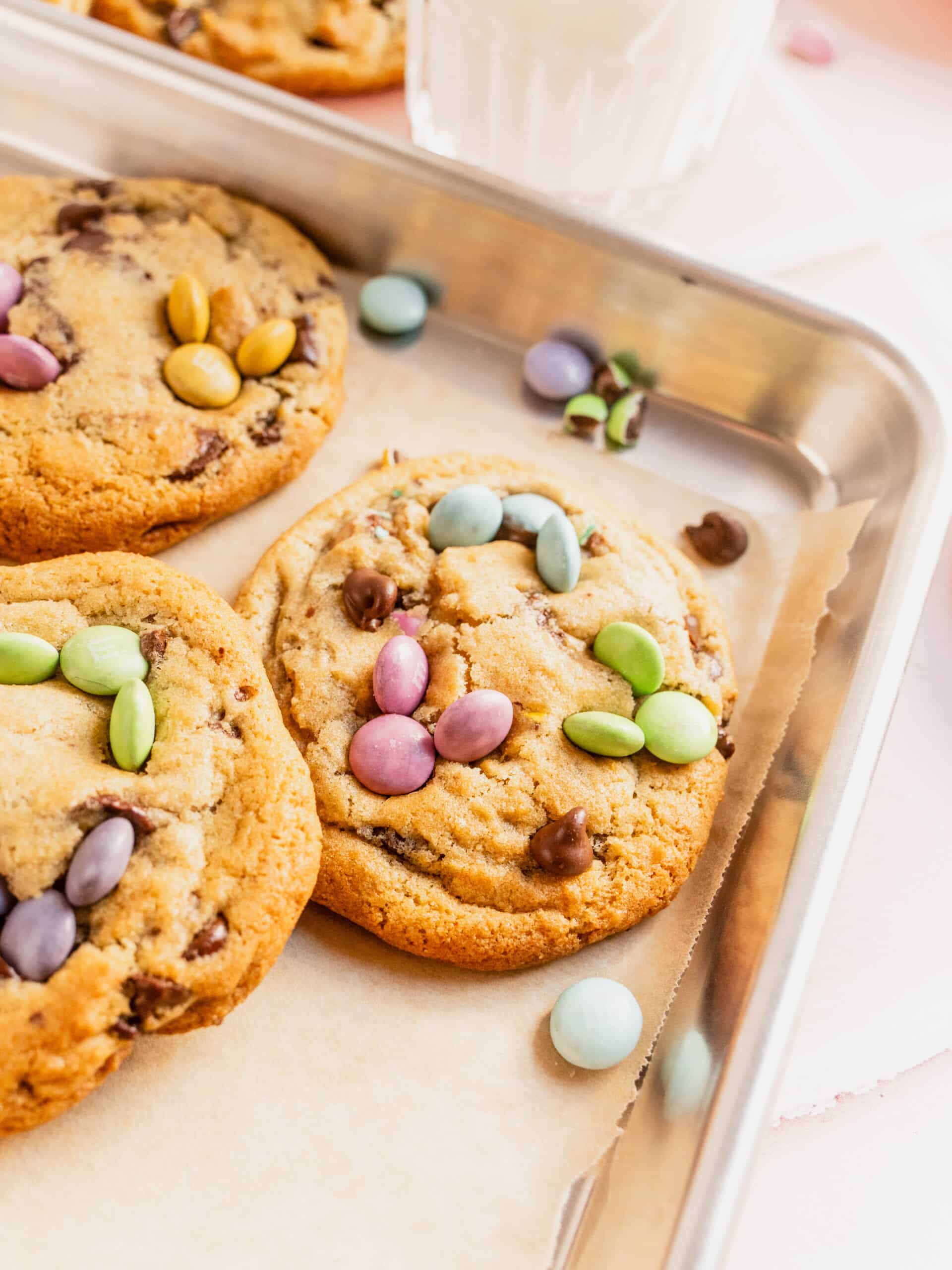 Close-up of freshly baked Easter bunny cookies on a parchment-lined baking sheet, topped with pastel M&Ms and chocolate chips, with golden edges and soft centers.