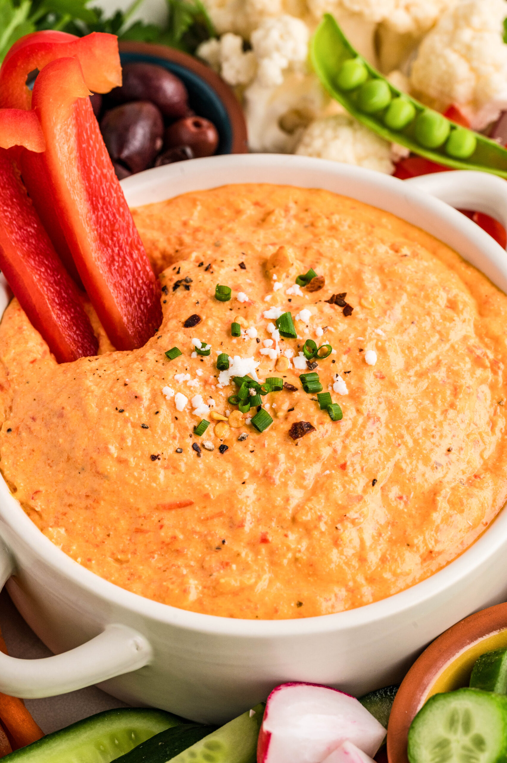 Close-up of creamy feta red pepper dip in a white serving bowl, topped with chopped chives, crumbled feta, cracked black pepper, and red pepper flakes. Fresh red bell pepper strips are dipped into the smooth, whipped dip, surrounded by cucumbers, radishes, olives, cauliflower, and sugar snap peas for serving.