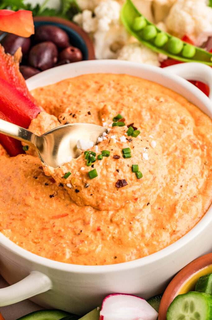 Close-up shot of creamy feta red pepper dip in a white serving bowl, topped with chopped chives, crumbled feta, and cracked black pepper. A spoon scoops into the smooth, whipped dip, surrounded by fresh vegetables including red bell pepper slices, cucumber, radishes, sugar snap peas, cauliflower, and a bowl of olives for dipping.