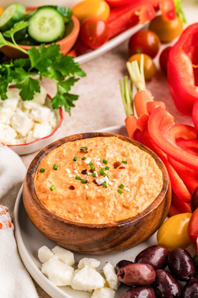 Close up shot of serving scene of creamy feta red pepper dip in a wooden bowl, topped with chopped chives, crumbled feta, and red pepper flakes. The dip is surrounded by fresh vegetables including red bell pepper slices, carrots, cucumbers, cherry tomatoes, cauliflower, olives, and parsley for dipping.