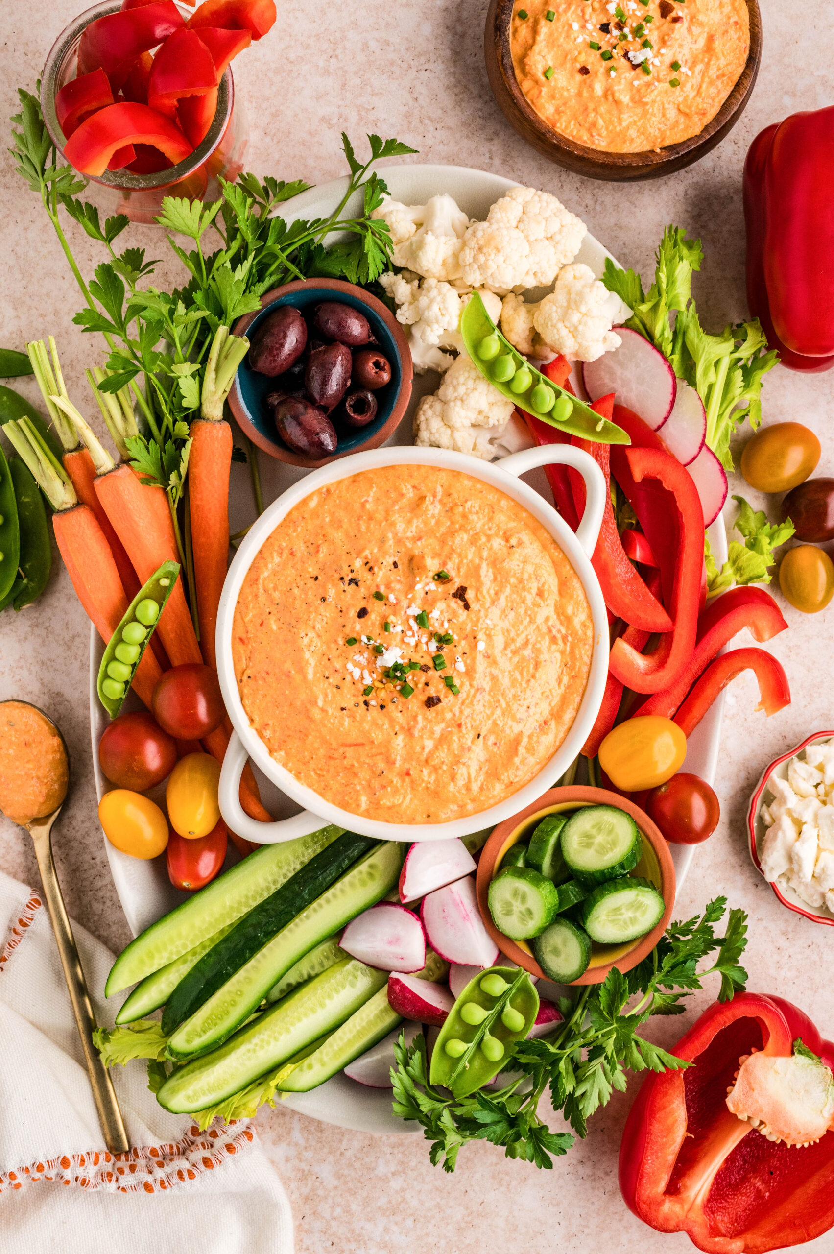 Overhead shot of creamy feta red pepper dip in a white serving bowl, topped with chopped chives, crumbled feta, and cracked black pepper. A spoon scoops into the smooth, whipped dip, surrounded by fresh vegetables including red bell pepper slices, cucumber, radishes, sugar snap peas, cauliflower, and a bowl of olives for dipping.