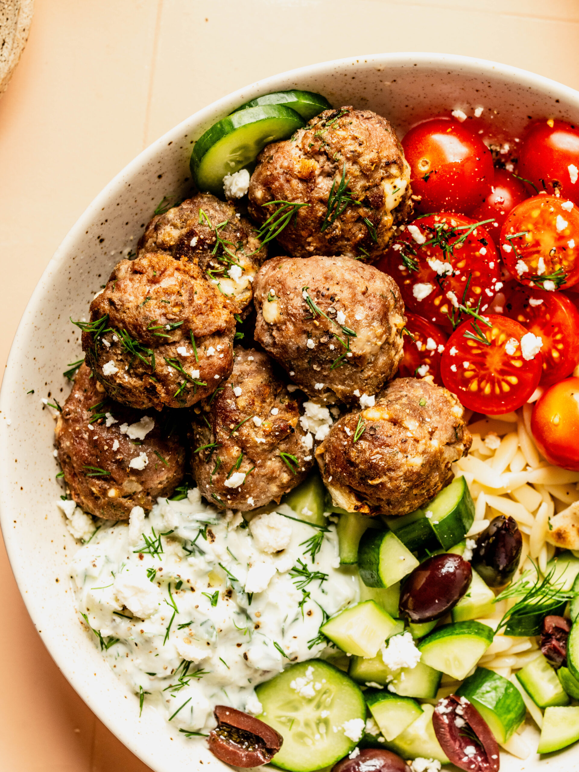 Mediterranean bowl with Greek feta meatballs served over orzo with cucumbers, cherry tomatoes, olives, and creamy tzatziki, finished with crumbled feta and fresh dill.