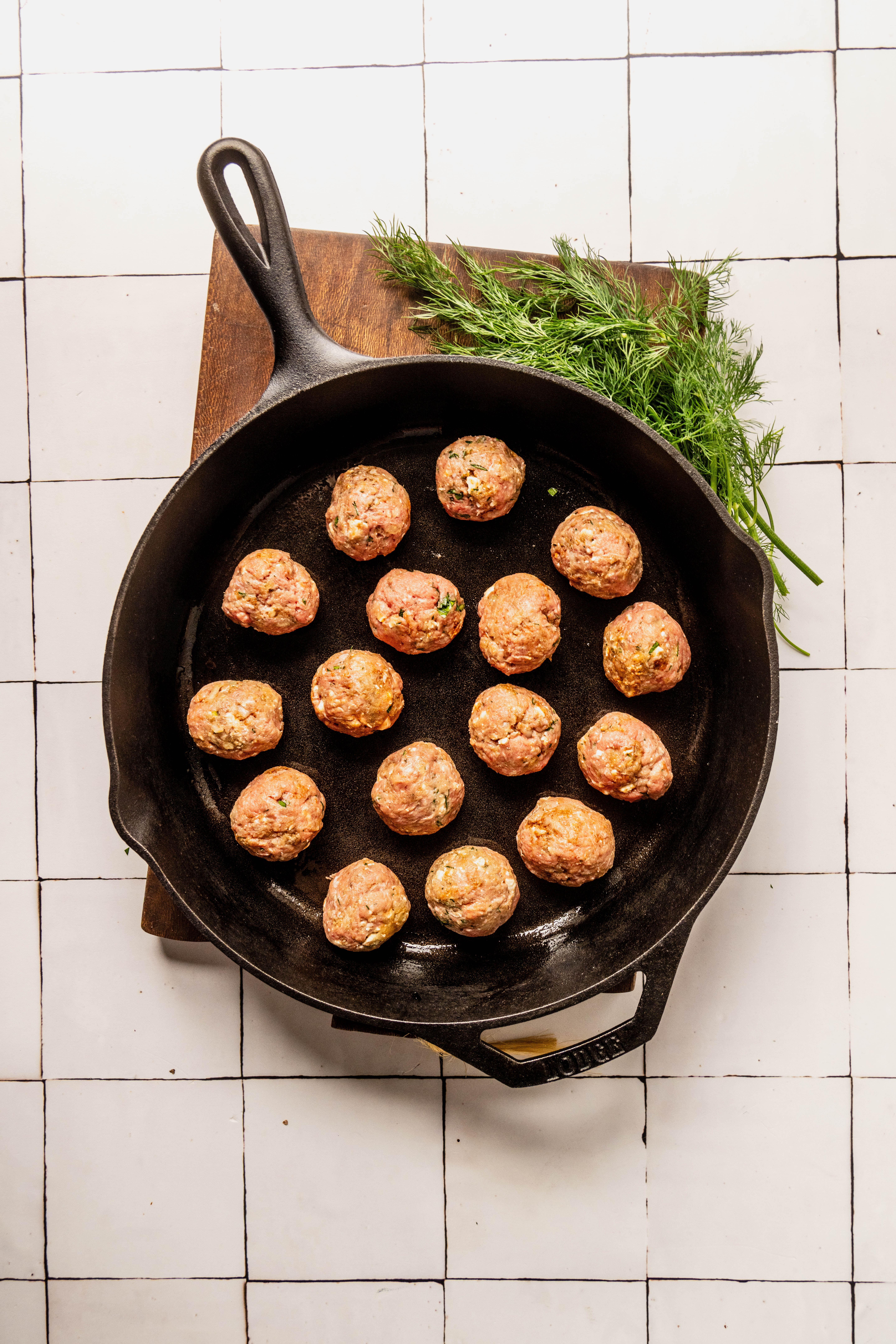 Raw Greek feta meatballs arranged in a cast iron skillet, ready to cook, with fresh dill on a wooden board over a white tiled background.