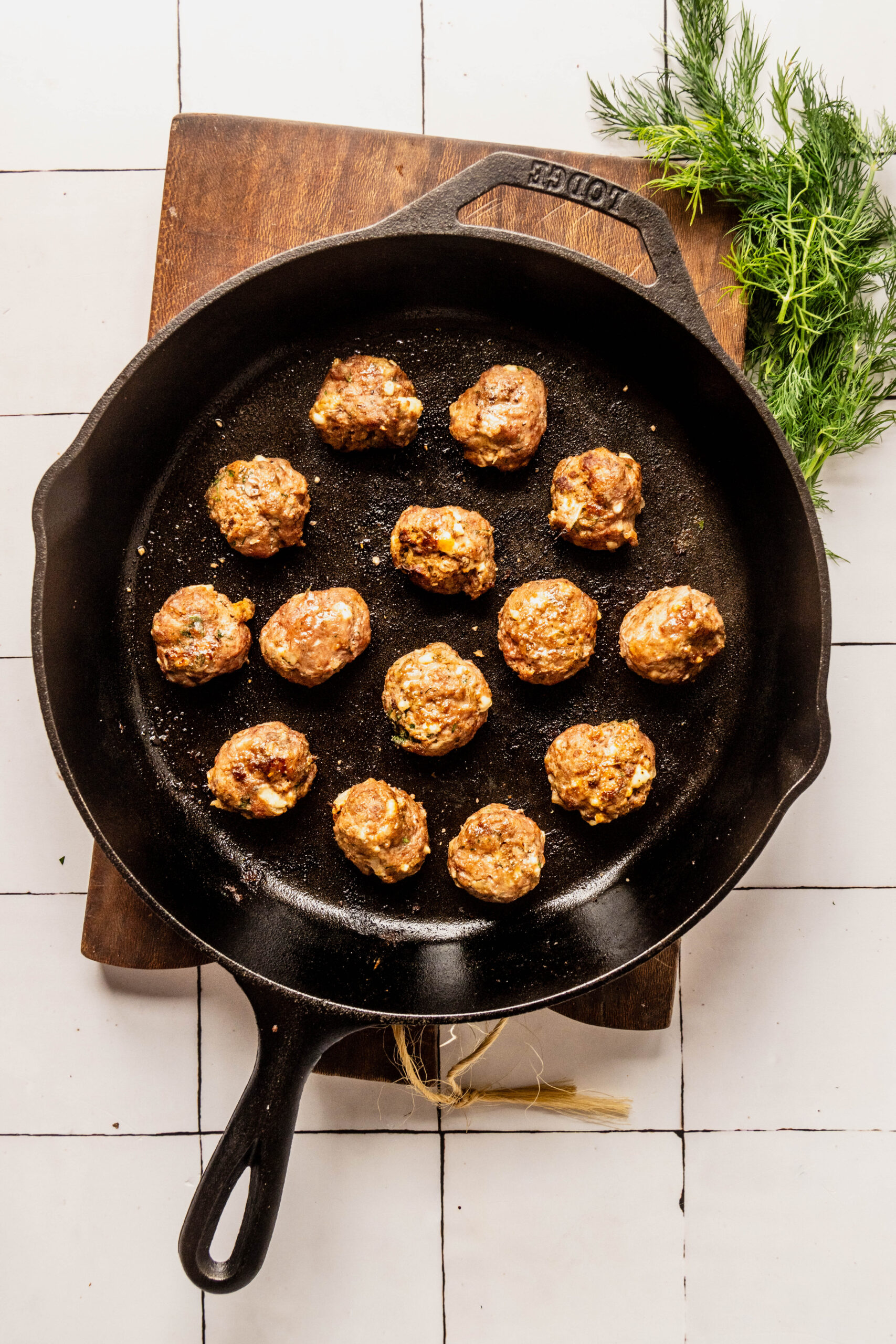 Cooked Greek feta meatballs arranged in a cast iron skillet, ready to cook, with fresh dill on a wooden board over a white tiled background.