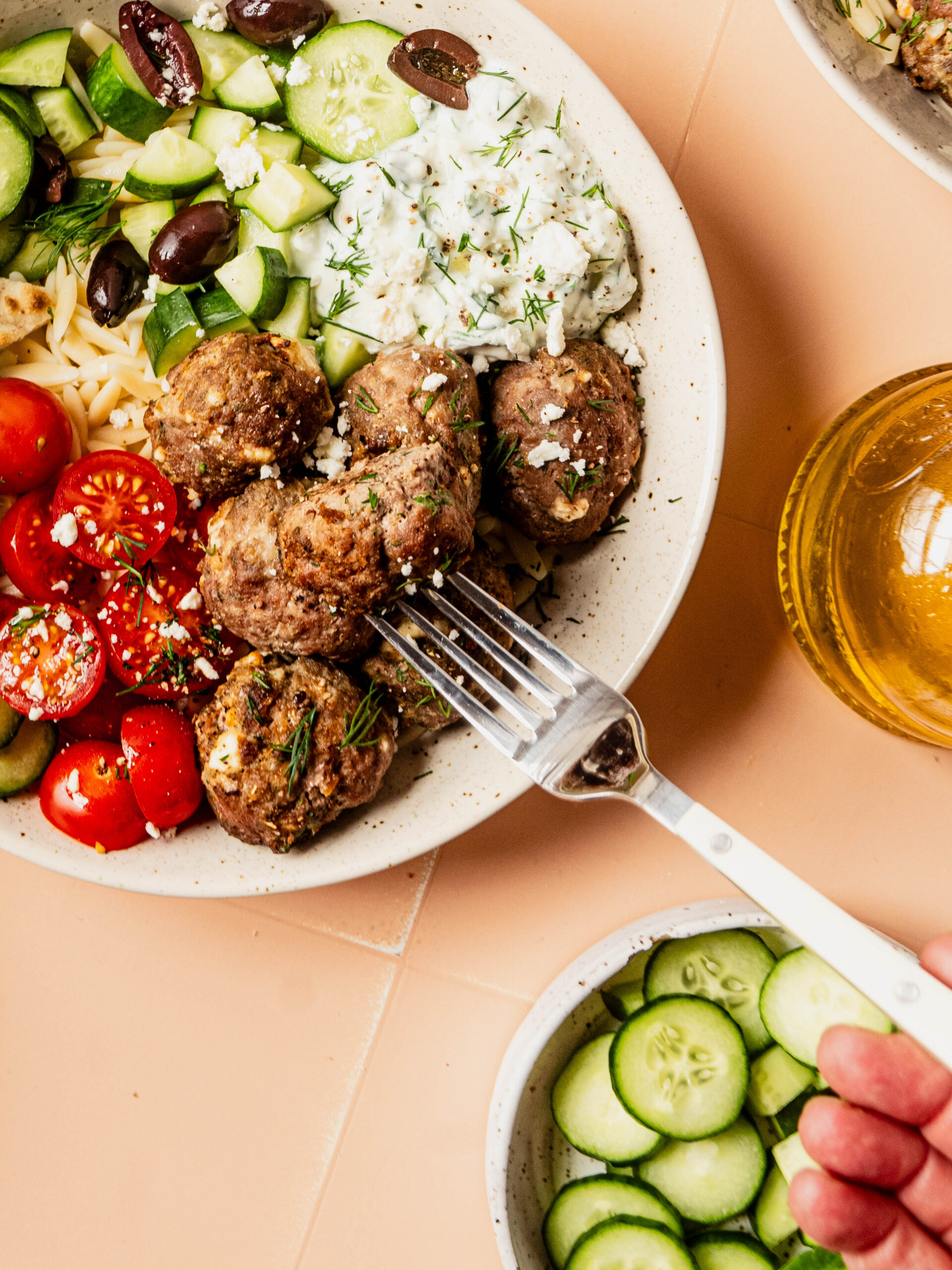 Fork lifting a Greek feta meatball from a Mediterranean bowl with orzo, cucumbers, cherry tomatoes, olives, and creamy tzatziki, topped with fresh dill and crumbled feta.