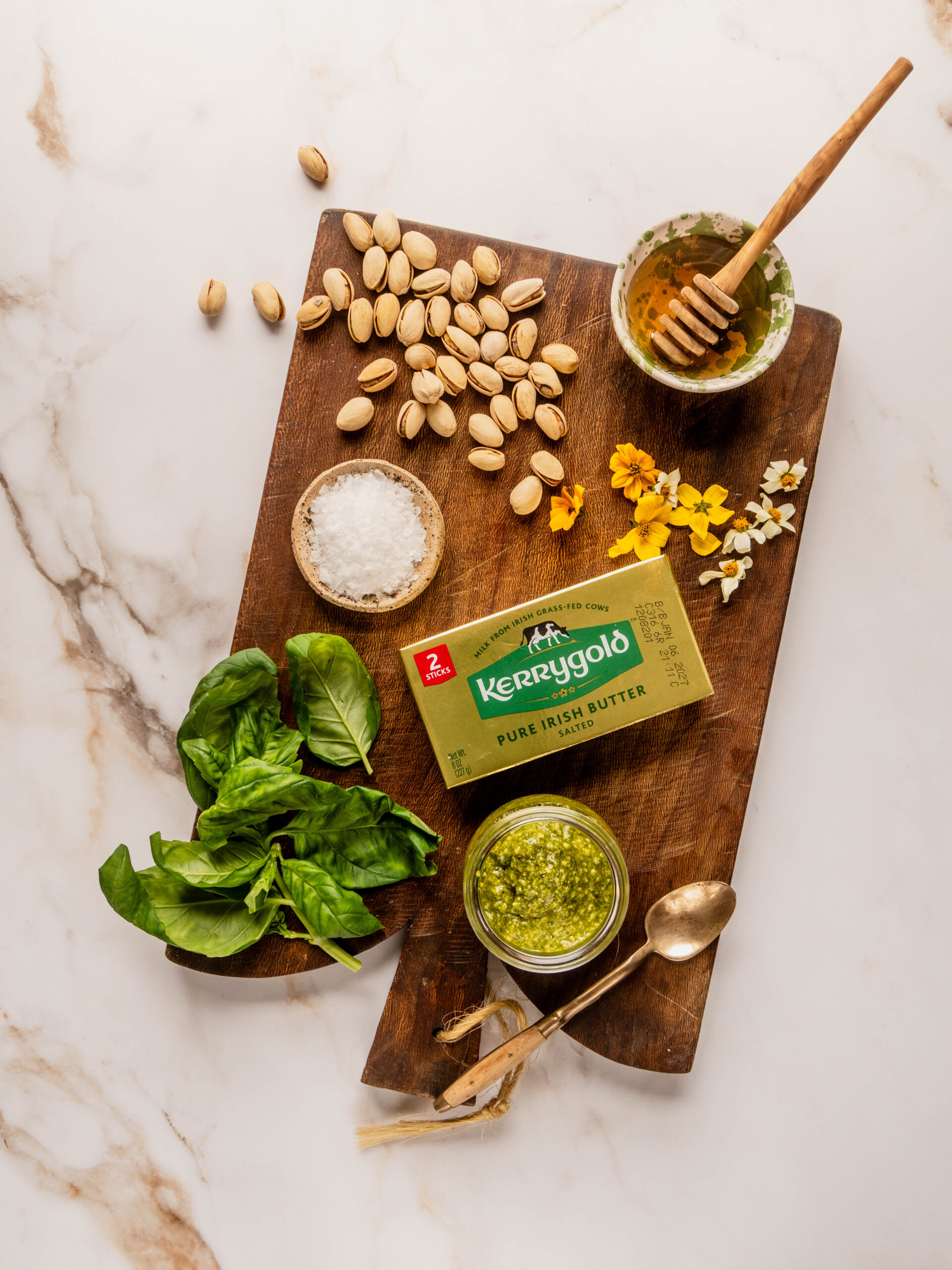 Ingredients for a pesto butter board arranged on a wooden board, including Kerrygold butter, fresh basil, pistachios, honey, pesto, flaky sea salt, and edible flowers on a marble background.