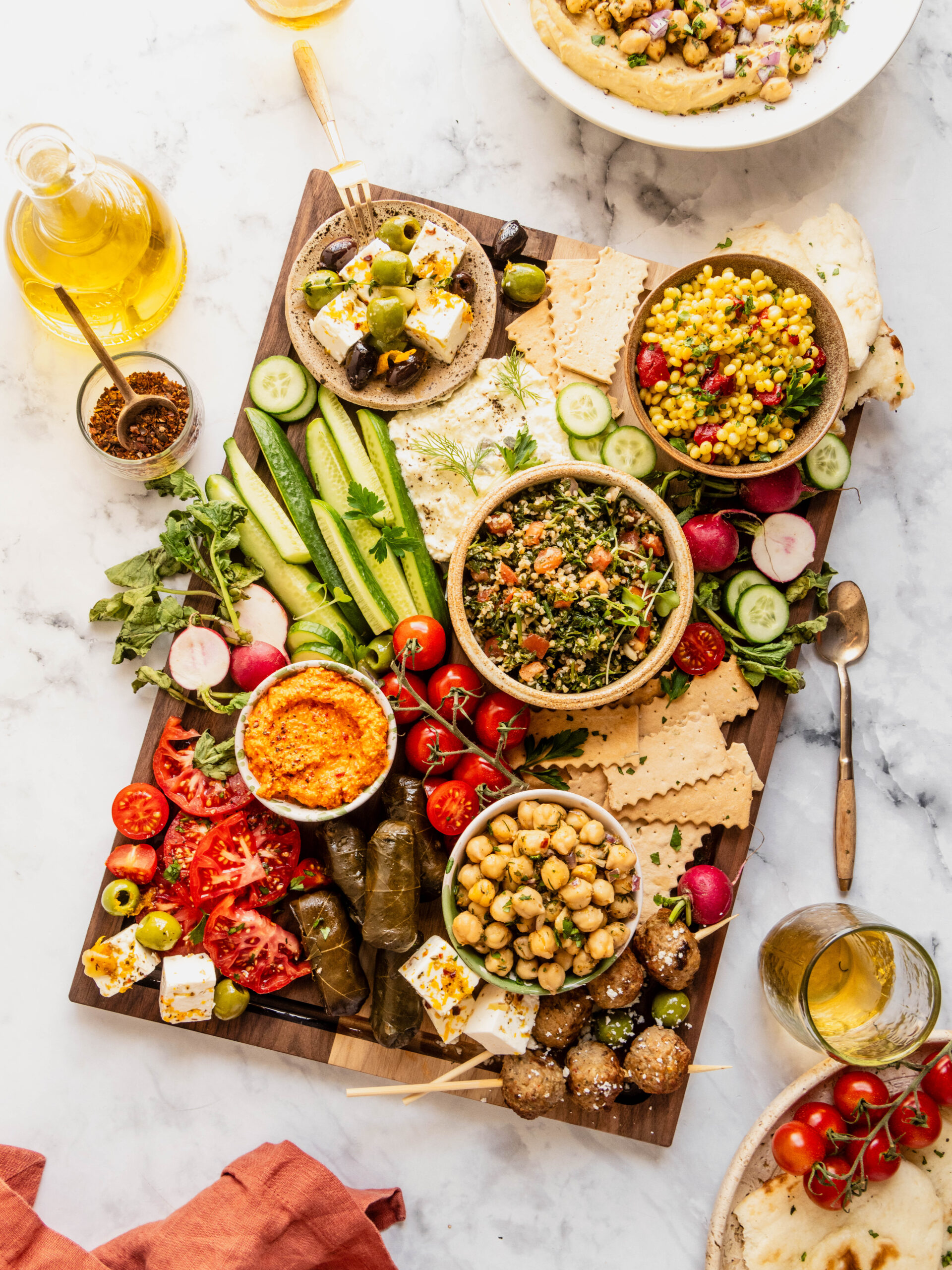 Overhead mezze board filled with colorful Mediterranean dishes including whipped feta, red pepper dip, tabouli, marinated chickpeas, feta and olives, grape leaves, fresh cucumbers, tomatoes, radishes, crackers, and Greek meatballs, styled on a wooden board with olive oil and wine on a marble background.