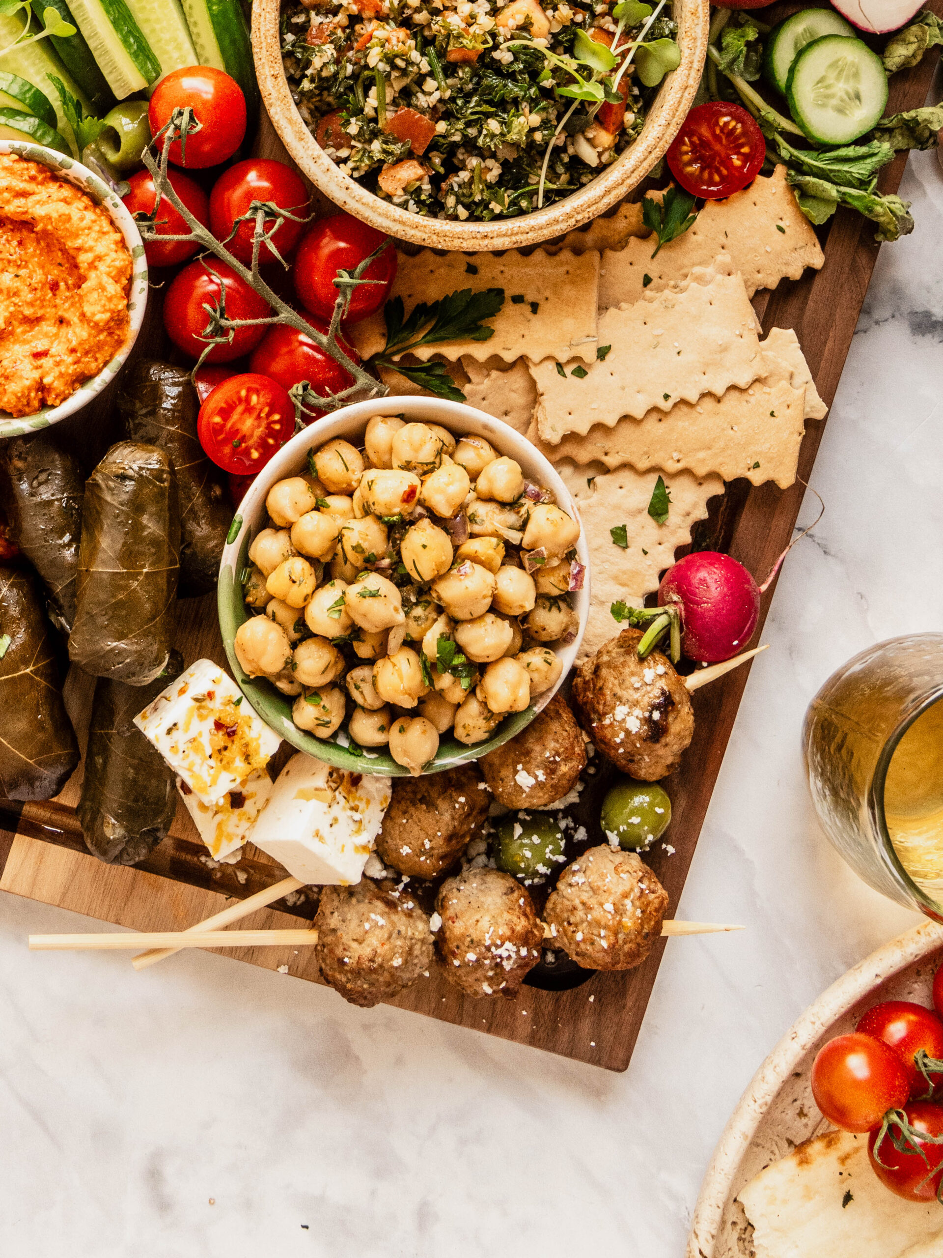Overhead close-up of a mezze board featuring marinated chickpeas, tabouli salad, red pepper dip, grape leaves, feta cubes, Greek meatballs on skewers, crackers, cherry tomatoes, cucumbers, and olives, styled on a wooden board with a bright, colorful presentation.