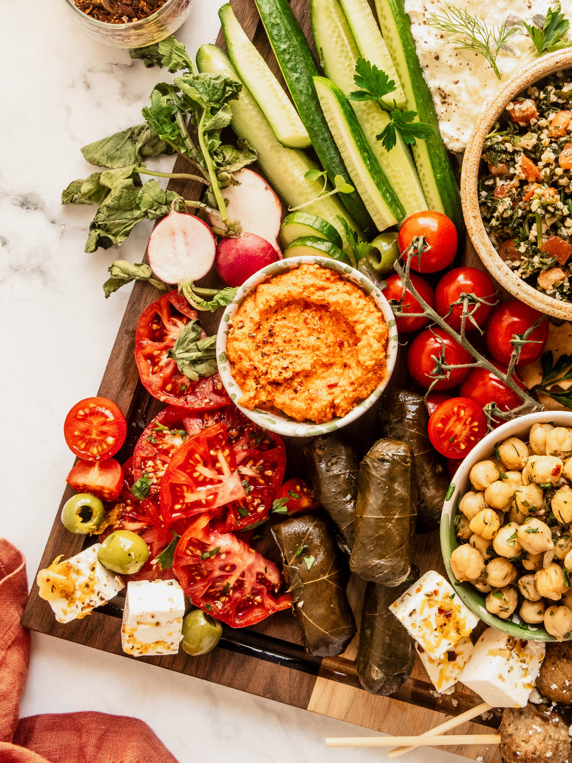 Colorful mezze board featuring red pepper dip, sliced heirloom tomatoes, cucumber spears, radishes, grape leaves, marinated chickpeas, feta cubes, olives, and fresh herbs, beautifully arranged on a wooden board with a bright, fresh overhead presentation.