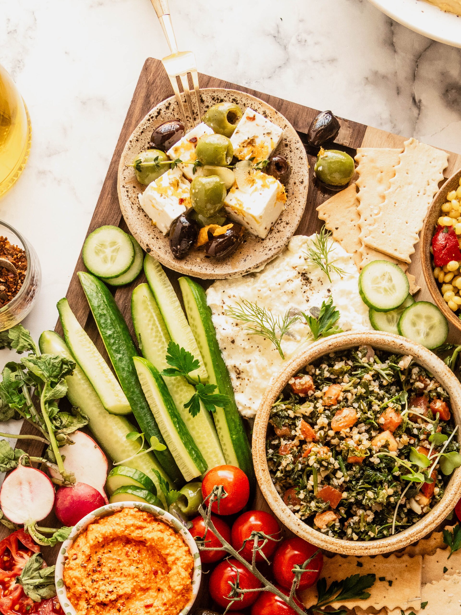 Bright, colorful mezze board styled on a wooden board with bowls of whipped feta and red pepper dip, tabouli salad, marinated olives and feta, fresh cucumber spears, radishes, cherry tomatoes, crackers, and herbs, photographed overhead on a marble surface with a light, airy aesthetic.