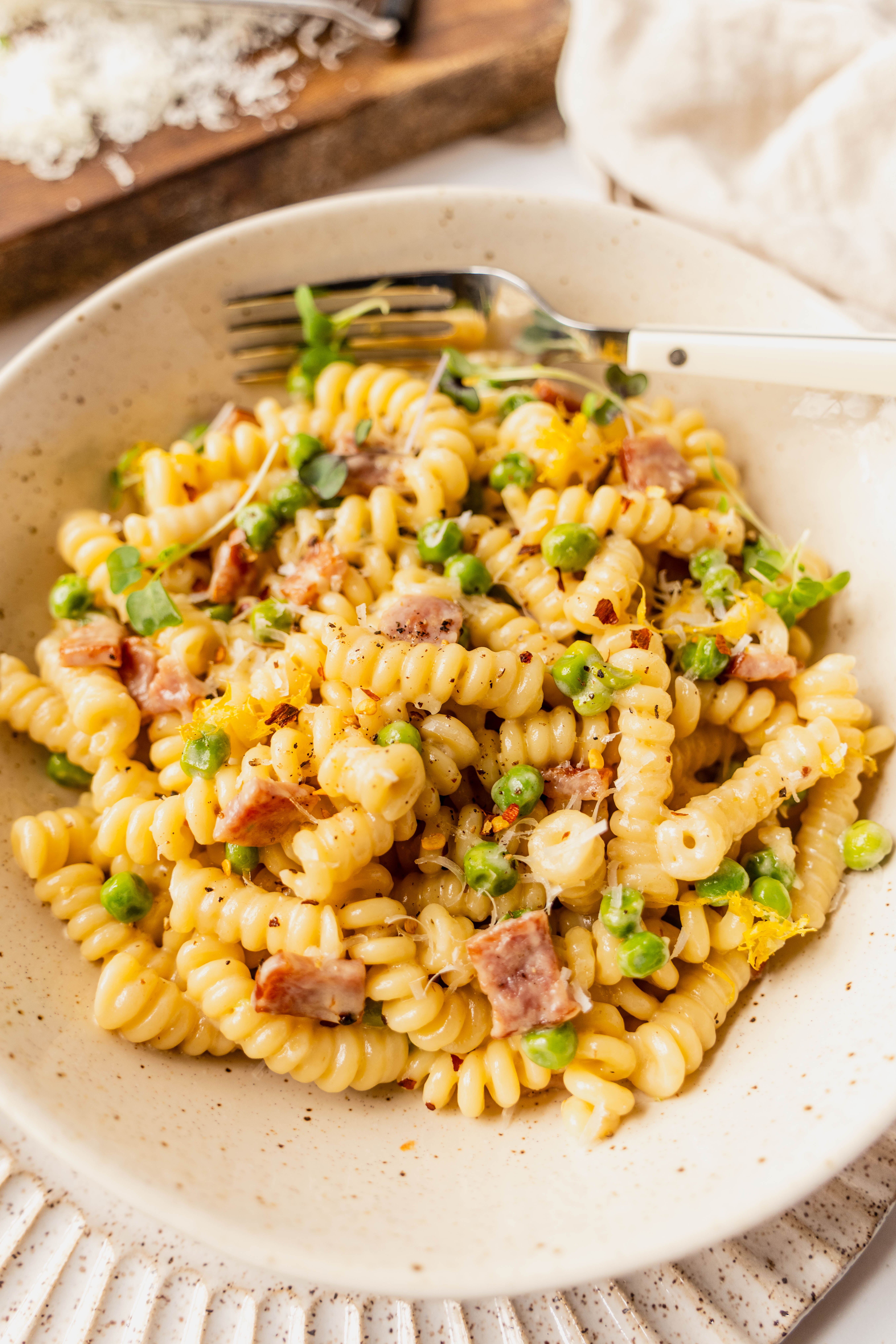 close-up of creamy pasta with peas and ham made with rotini noodles tossed in a silky Parmesan cream sauce, dotted with bright green peas and golden cubes of ham, finished with freshly grated cheese, cracked black pepper, red pepper flakes, and lemon zest in a neutral ceramic bowl.