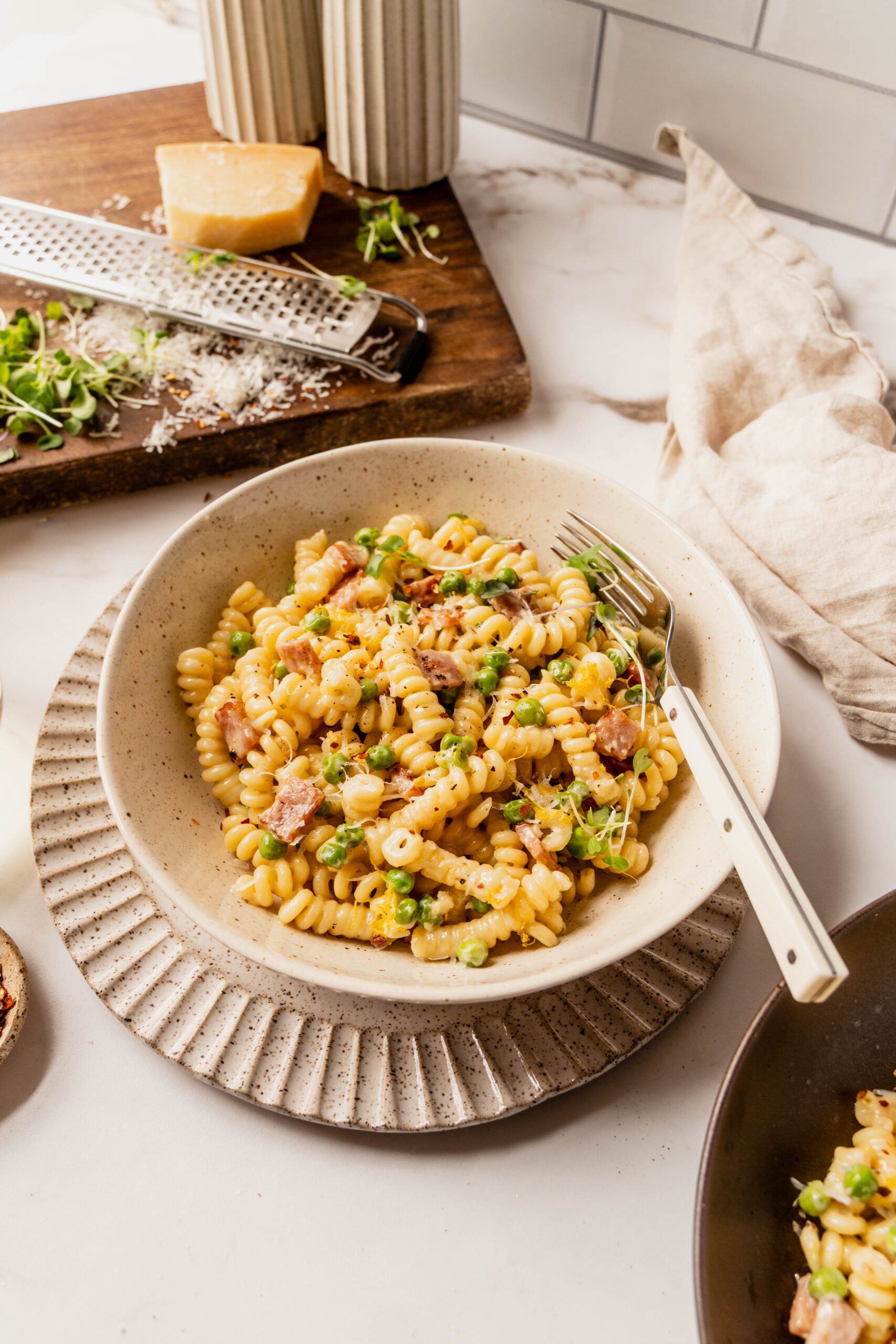 45 degree angle shot of a kitchen scene featuring pasta with peas and ham made with rotini in a creamy Parmesan sauce, topped with lemon zest, cracked black pepper, and red pepper flakes, served in a speckled ceramic bowl with a fork, alongside a block of Parmesan and grater on a wooden board.