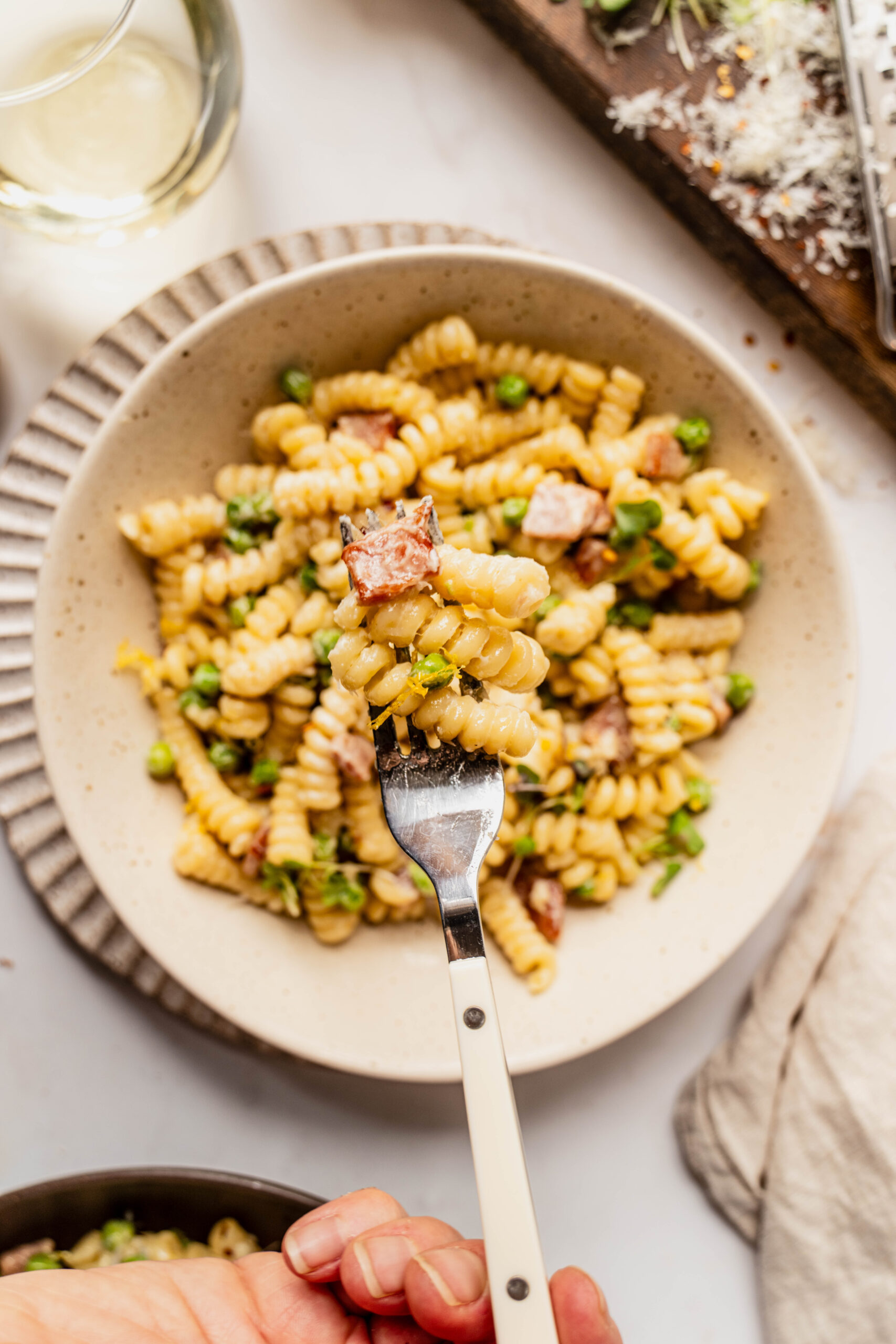 Close-up overhead view of pasta with peas and ham featuring creamy rotini coated in Parmesan sauce, mixed with bright green peas and golden cubes of ham, as a fork lifts a bite topped with lemon zest and cracked black pepper from a neutral ceramic bowl.