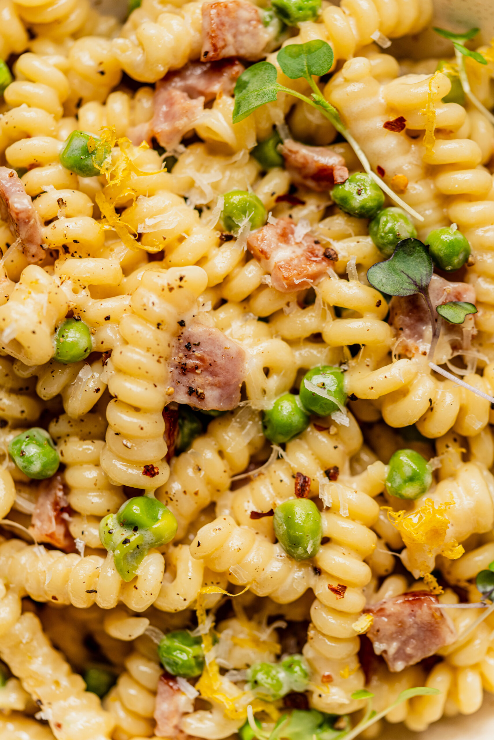 Macro shot of pasta with peas and ham made with rotini in a creamy Parmesan sauce, topped with lemon zest, cracked black pepper, and red pepper flakes