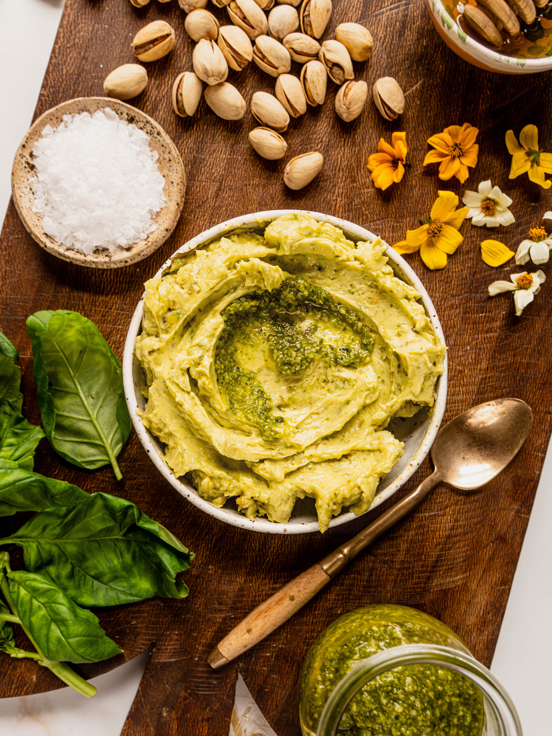 Bowl of pesto butter for a butter board surrounded by fresh basil, pistachios, flaky sea salt, edible flowers, and a jar of pesto on a wooden board.