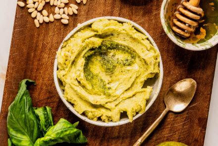 Overhead view of creamy pesto butter in a small bowl on a wooden board surrounded by fresh basil leaves, pine nuts, flaky sea salt, honey with a wooden dipper, and a jar of pesto on a white marble background.