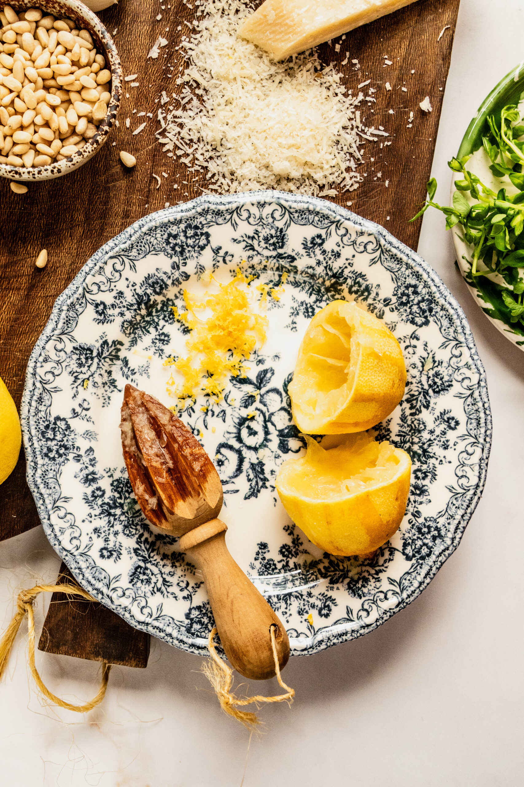 Alt text: Overhead shot of fresh lemon and grated zest on a patterned plate with a wooden citrus reamer, alongside Parmesan and pine nuts on a cutting board, ingredients for bright pesto goat cheese pasta.