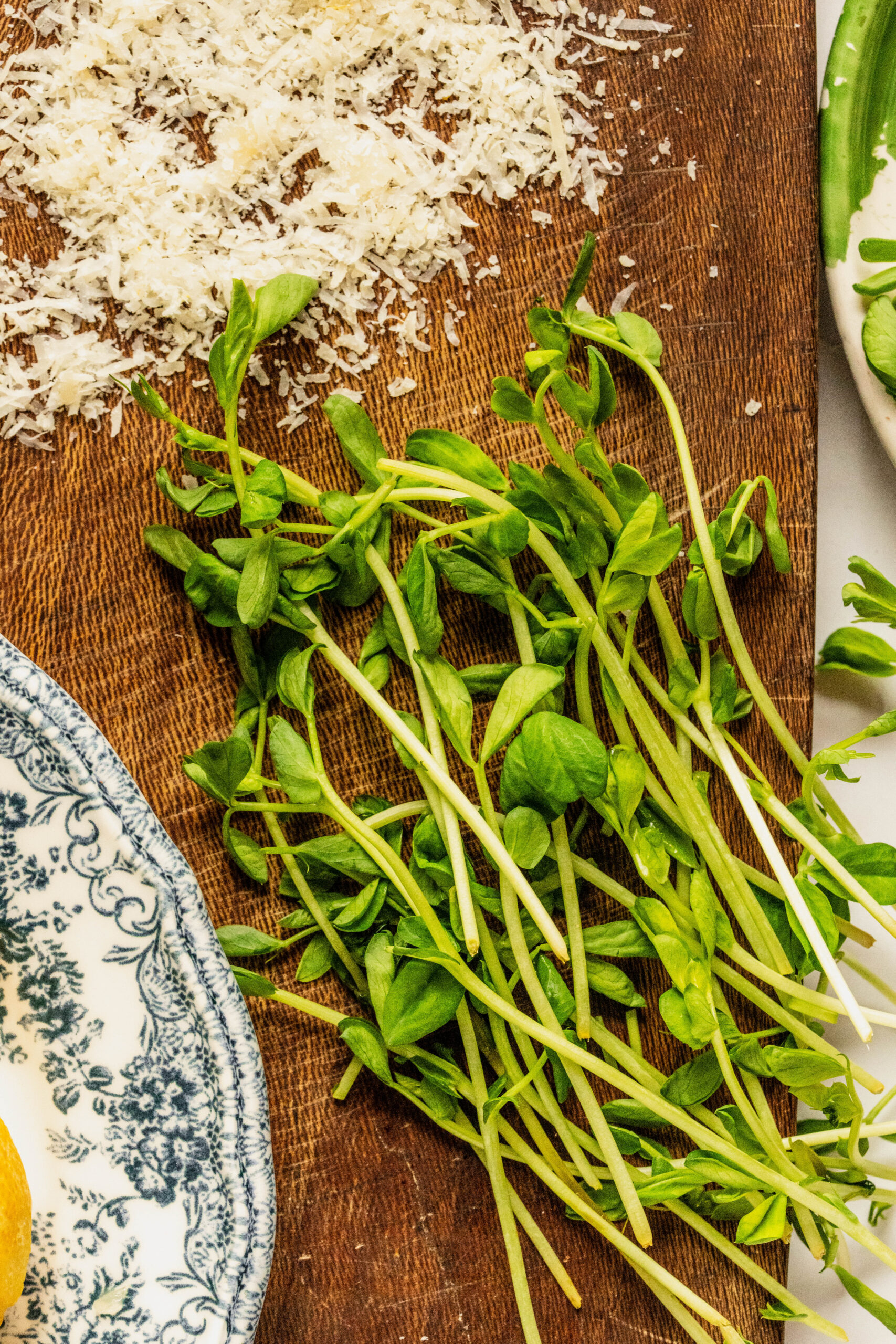 Close-up of fresh greens and grated cheese on a wooden board for pesto goat cheese pasta, featuring tender arugula or pea shoots alongside a pile of finely grated Parmesan, styled for a bright, fresh pasta preparation.