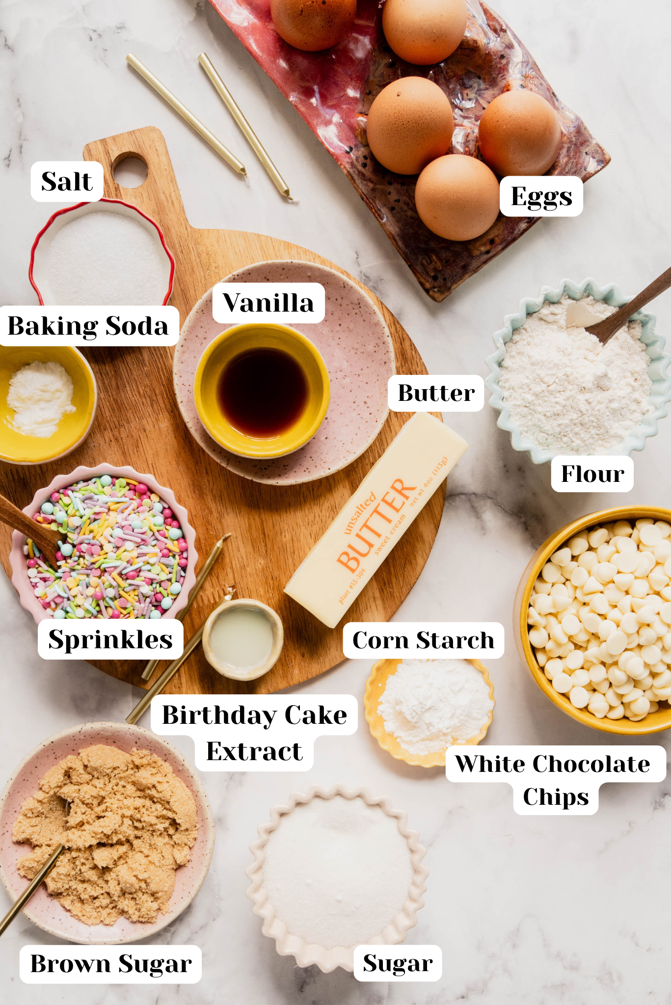 Bright overhead flat lay of ingredients for birthday cake cookies arranged on a white marble surface, including flour, butter, eggs, sugar, brown sugar, vanilla, birthday cake extract, cornstarch, baking soda, salt, white chocolate chips, and colorful sprinkles in small bowls and dishes.