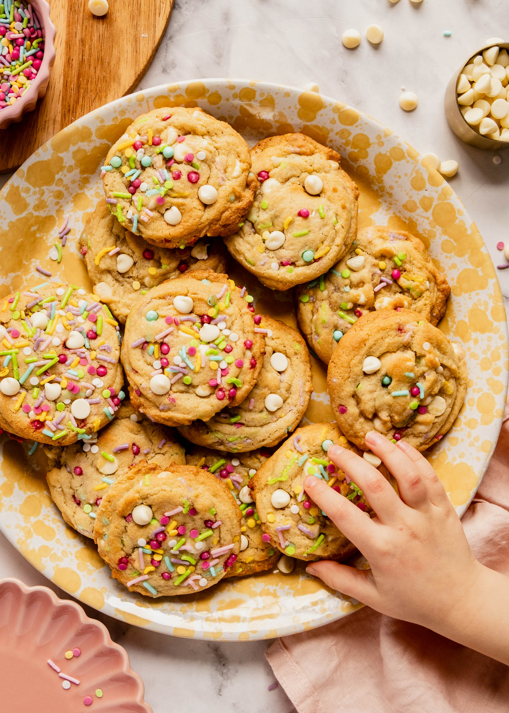 Bright overhead image of soft birthday cake cookies topped with colorful sprinkles and white chocolate chips on a speckled plate, with a hand reaching in to grab one, surrounded by extra sprinkles and baking ingredients on a marble surface.