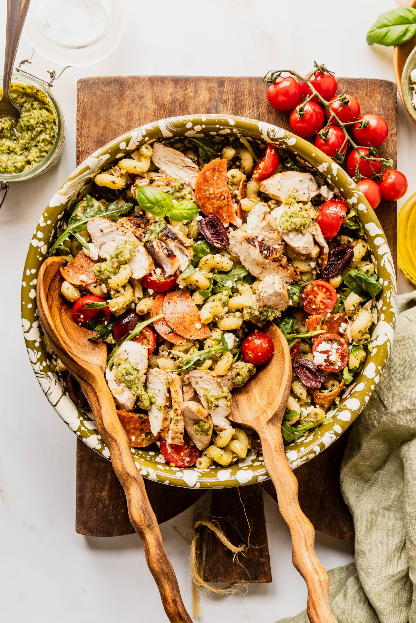 Overhead view of pasta salad pesto chicken in a large serving bowl with corkscrew pasta tossed in basil pesto, grilled chicken slices, cherry tomatoes, kalamata olives, pepperoni, feta cheese, and arugula, served with wooden salad spoons on a marble surface.
